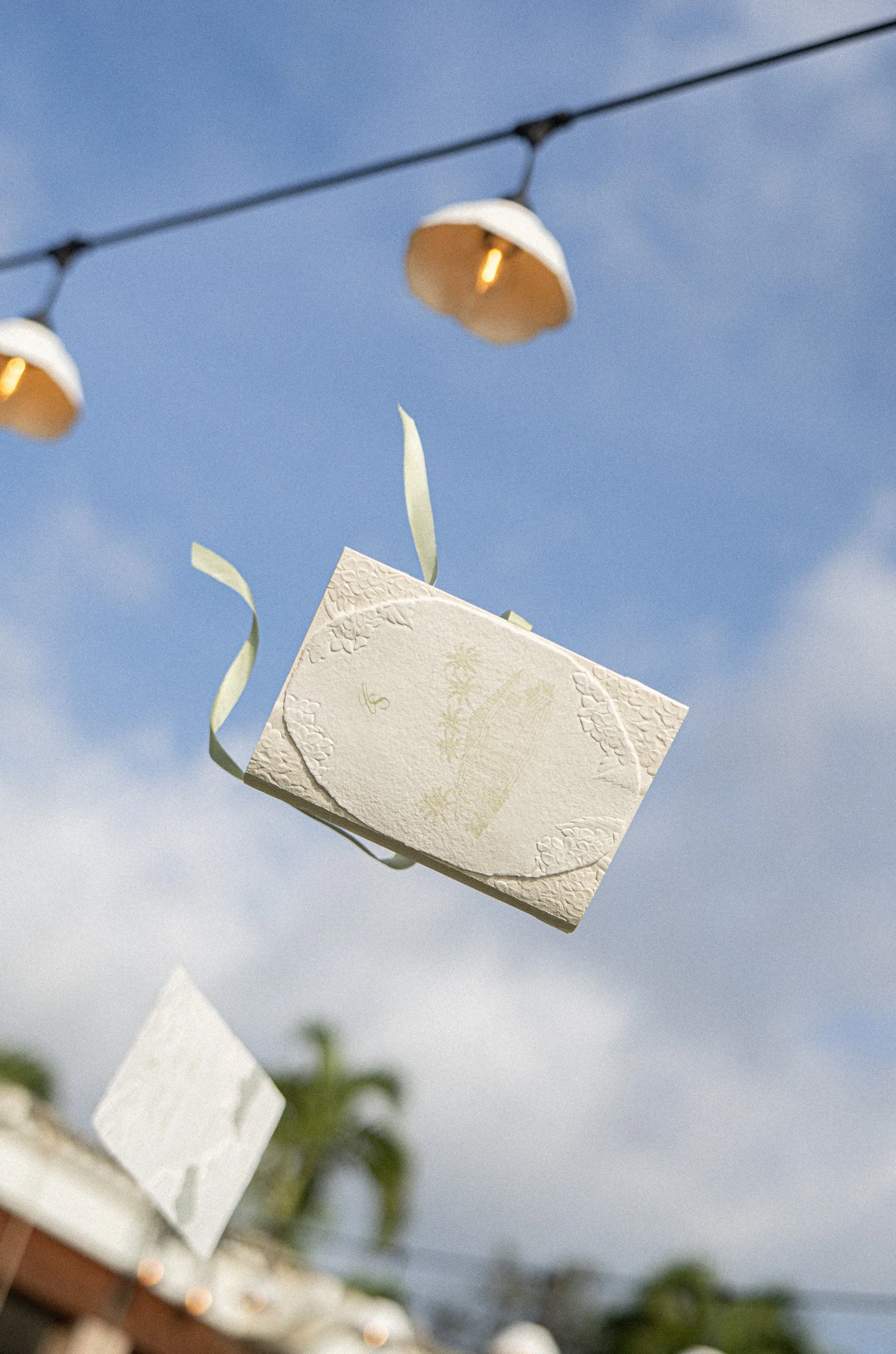 String lights hanging outdoors against a blue sky, with a white textured envelope or card in the foreground.