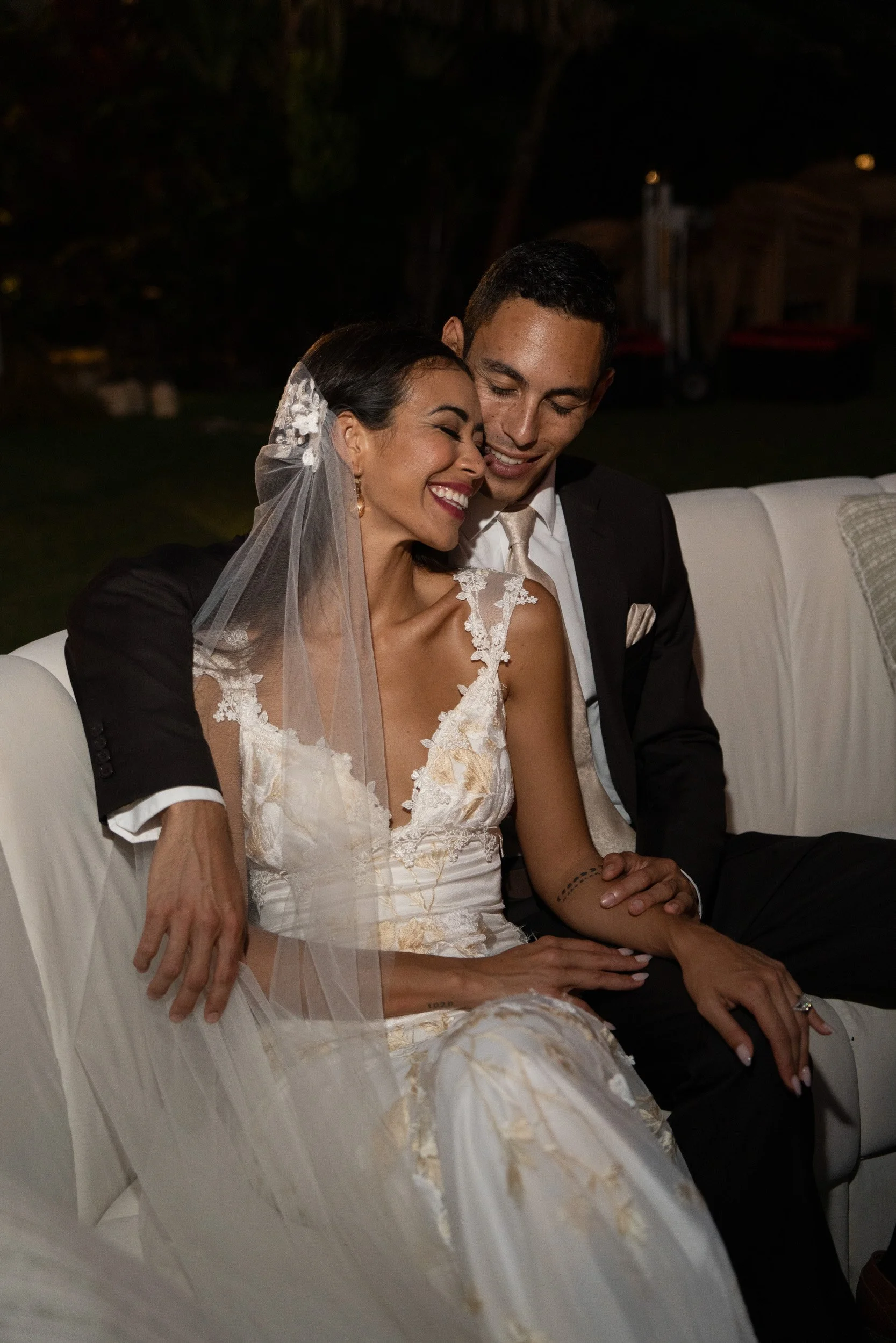 A bride and groom sitting closely on a white sofa, smiling and enjoying a moment together during their wedding reception at night, with the bride wearing a lace wedding dress and veil, and the groom in a dark suit.