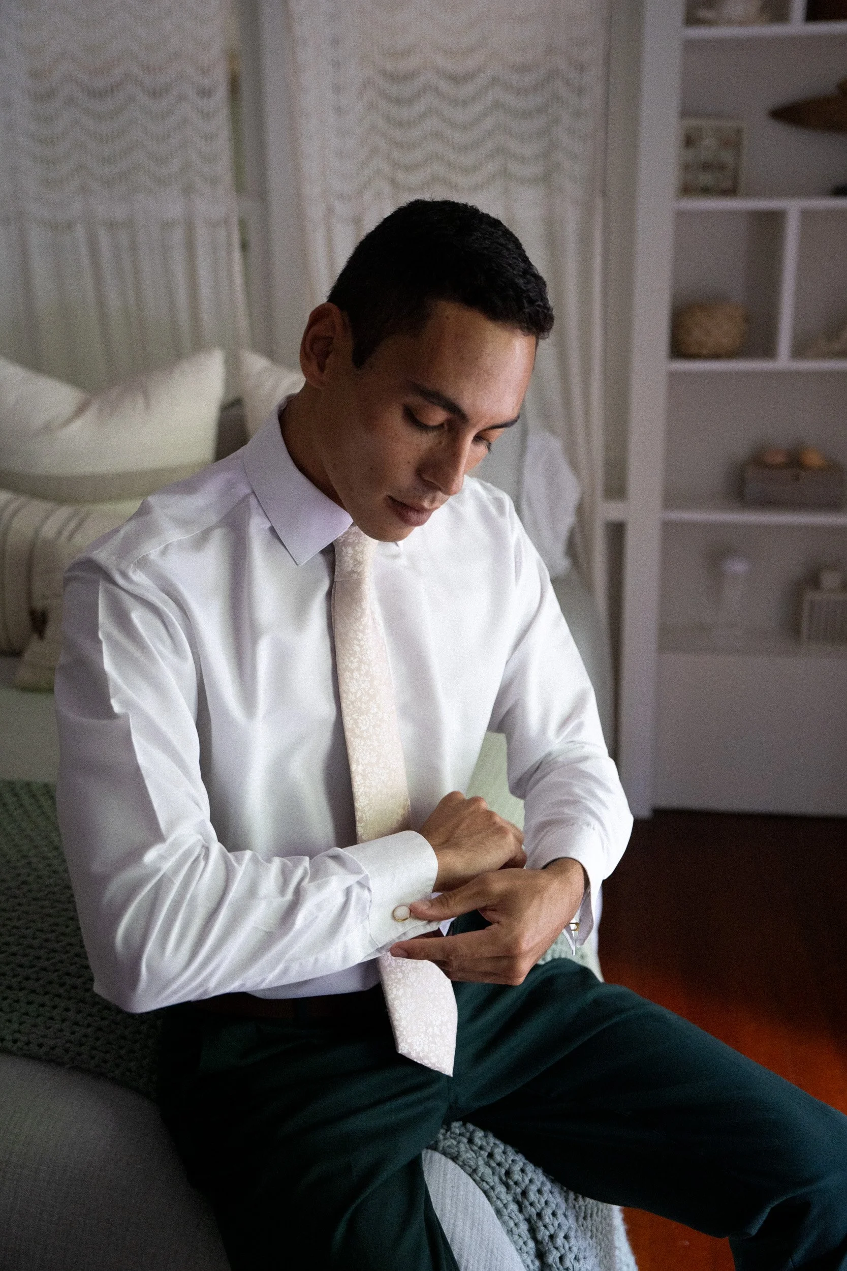 A man sitting on a gray couch, putting on a watch, wearing a white dress shirt and a silk tie, in a room with shelves and white curtains.