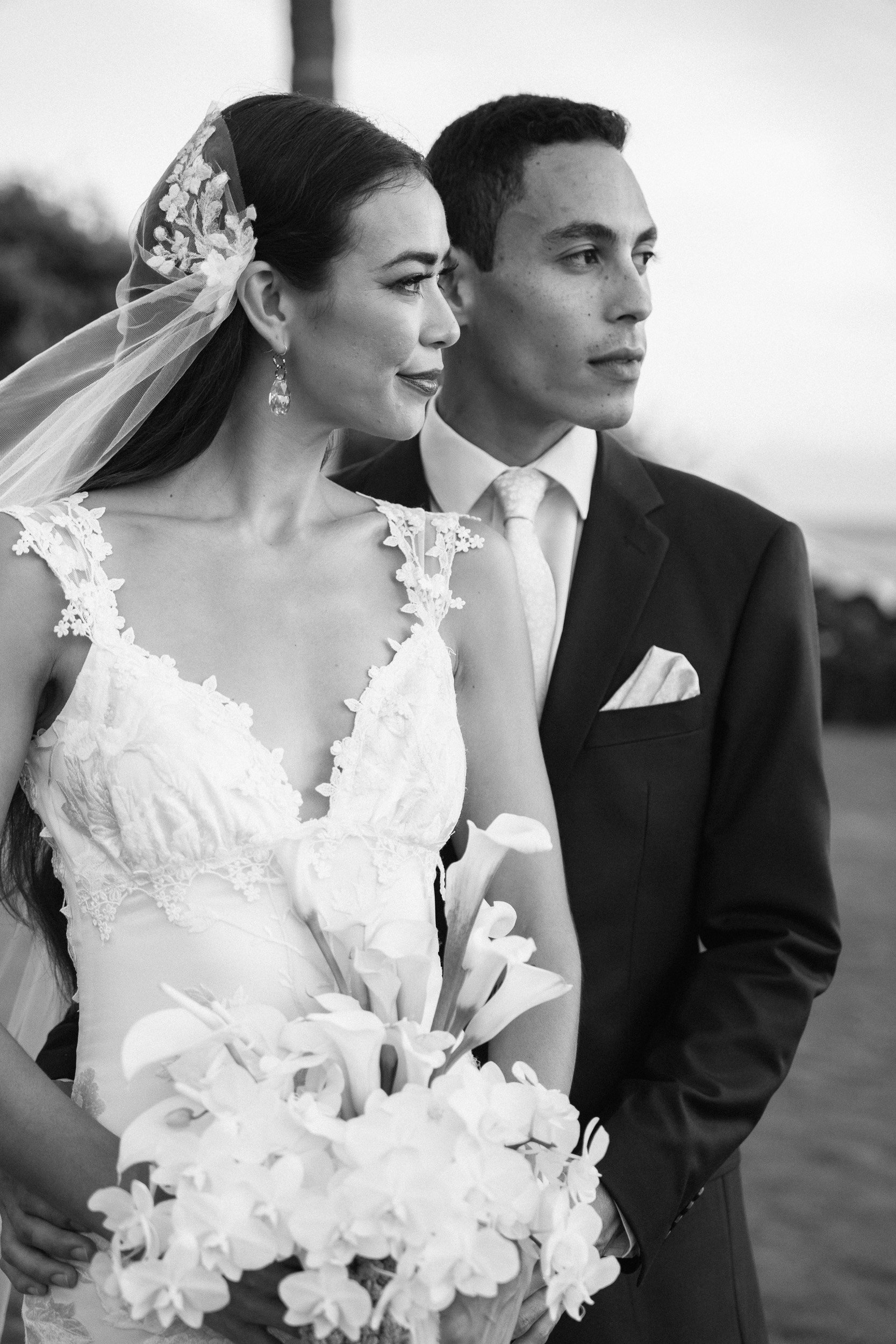 A black and white photo of a bride and groom at their wedding, standing side by side outdoors. The bride wears a lace wedding dress with a veil and earrings, holding a bouquet of flowers. The groom is dressed in a dark suit with a white shirt and tie