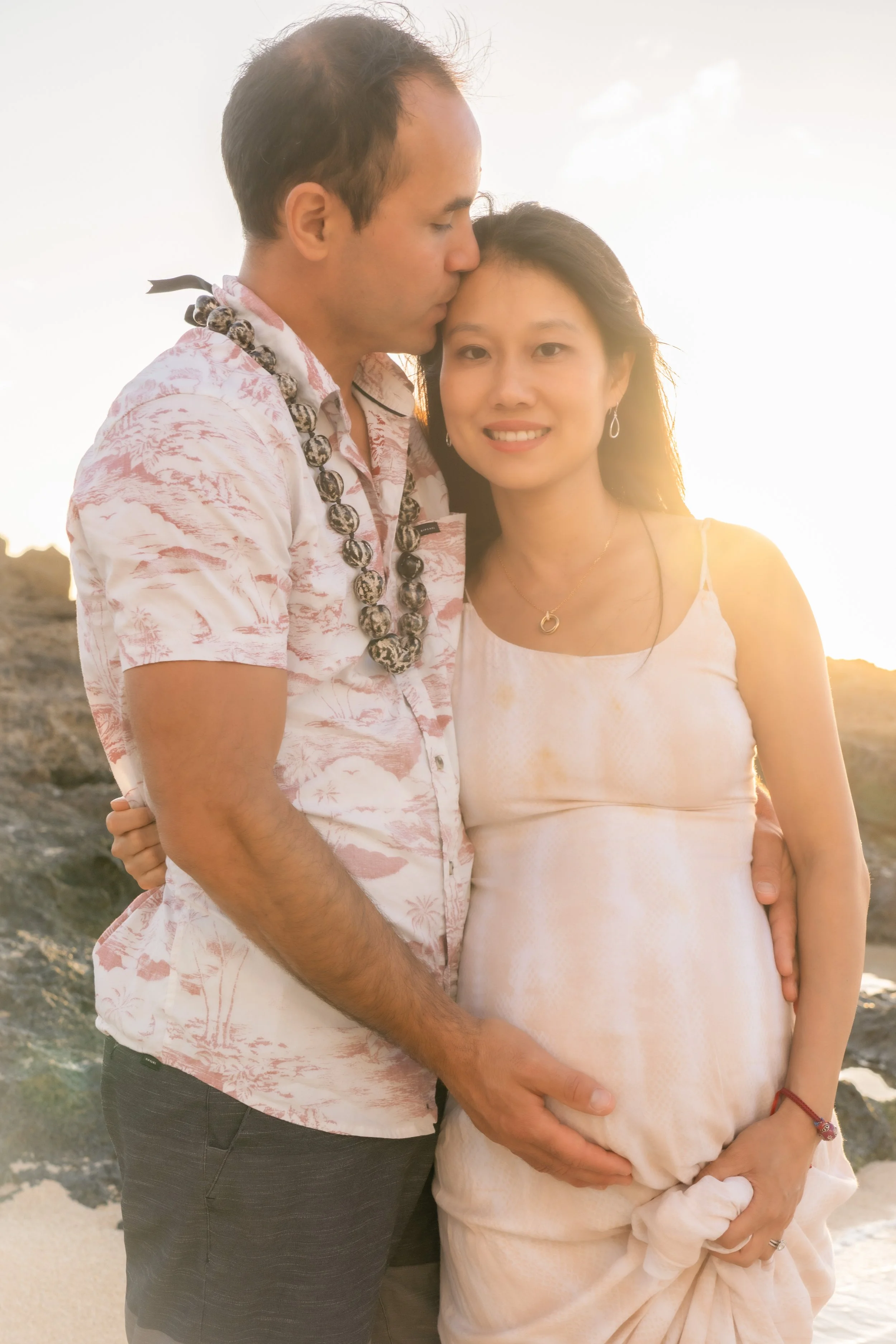 A pregnant woman with long black hair and earrings, and a man with short dark hair, stand close together on a beach during sunset, with rocks visible behind them. The man, wearing a patterned short-sleeve shirt and beaded necklace, kisses the woman's