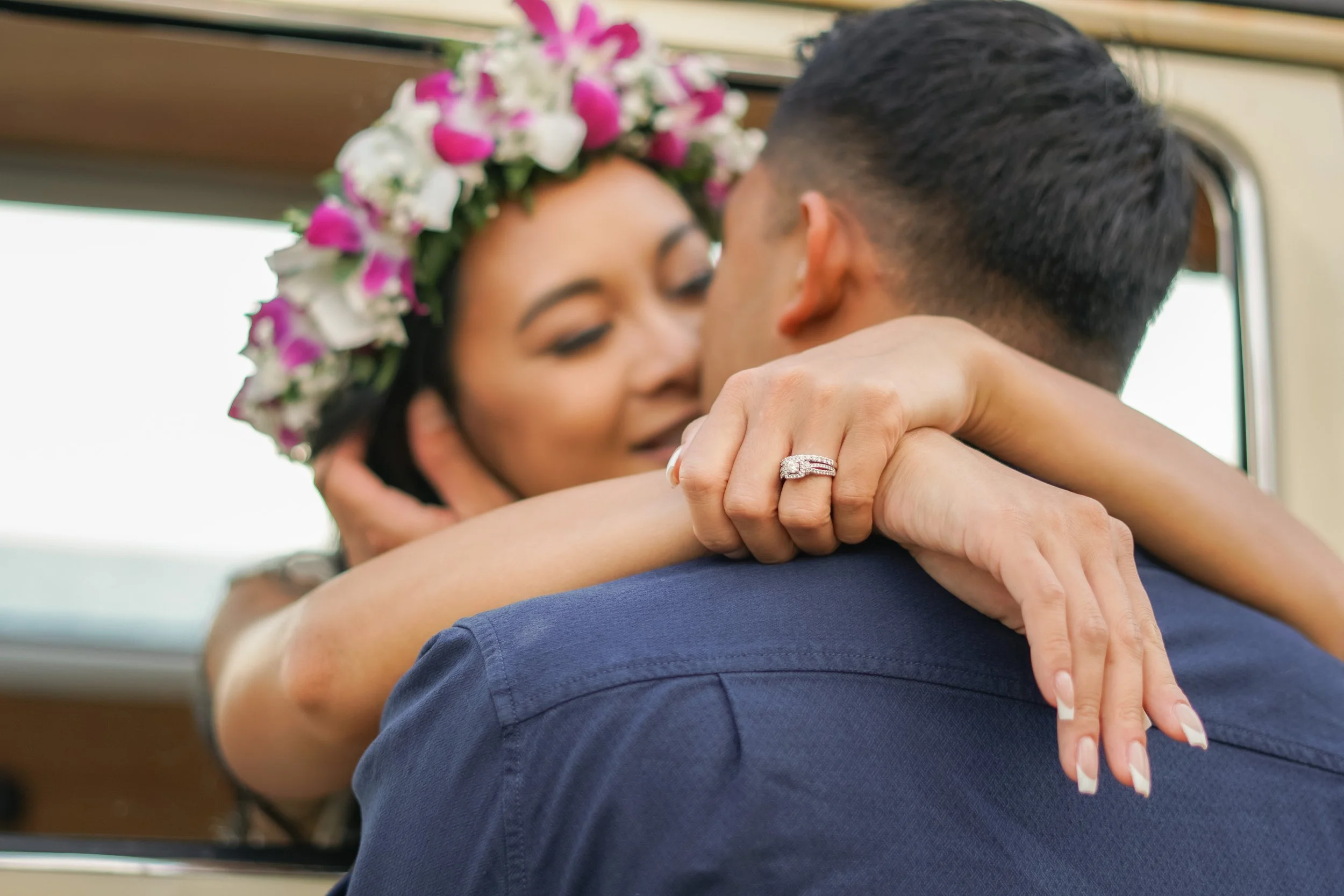 A woman wearing a floral crown and an engagement ring is hugging a man inside a vehicle, their faces close together.