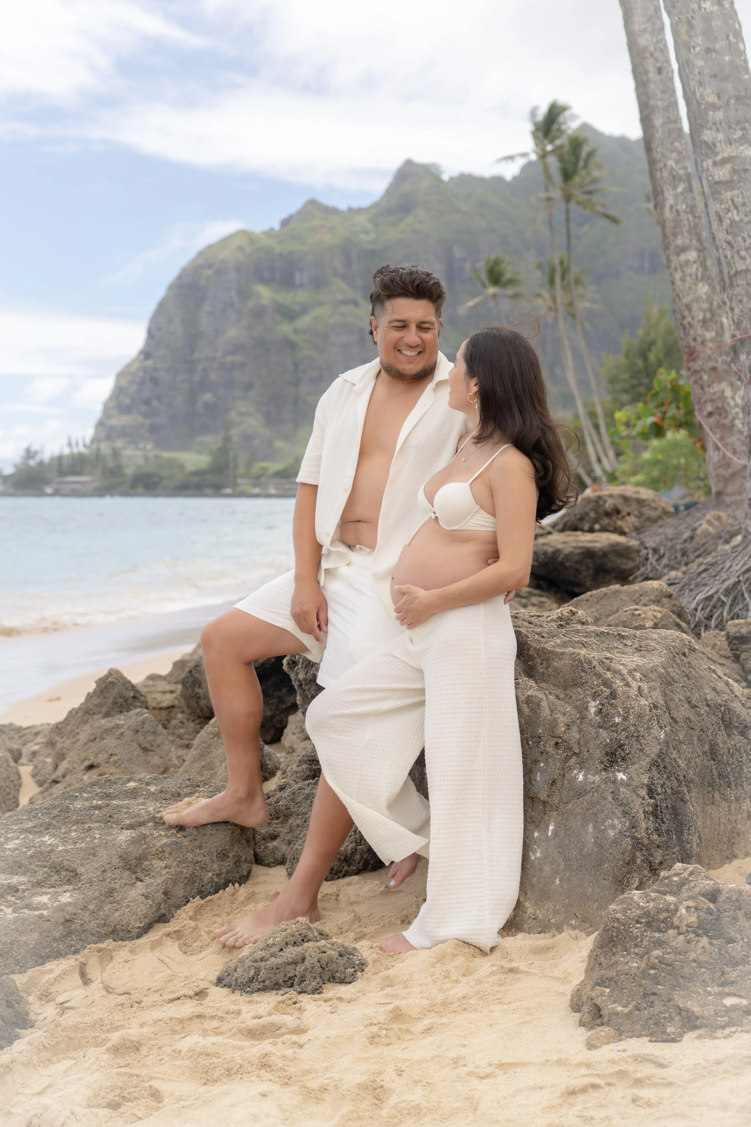 A pregnant woman and a man are standing on a rocky beach, with the woman leaning against a large rock. They are dressed in light, casual clothing and are smiling at each other. The background features a mountain, palm trees, and the ocean under a partly cloudy sky.