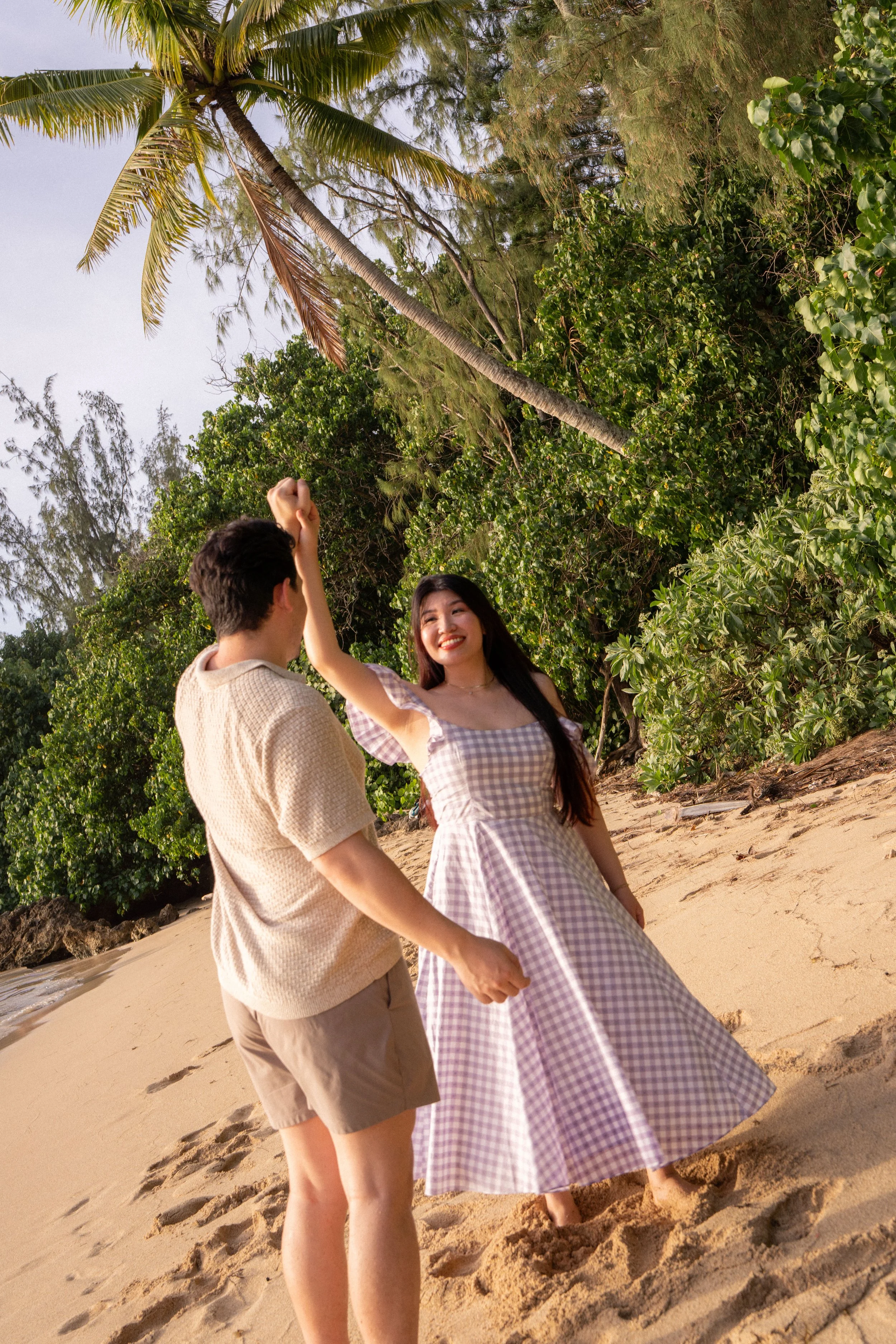 A man and woman on a sandy beach with lush green trees and palm trees in the background. The man is wearing a beige shirt and shorts, and the woman is wearing a purple checkered dress. They are holding hands and smiling.
