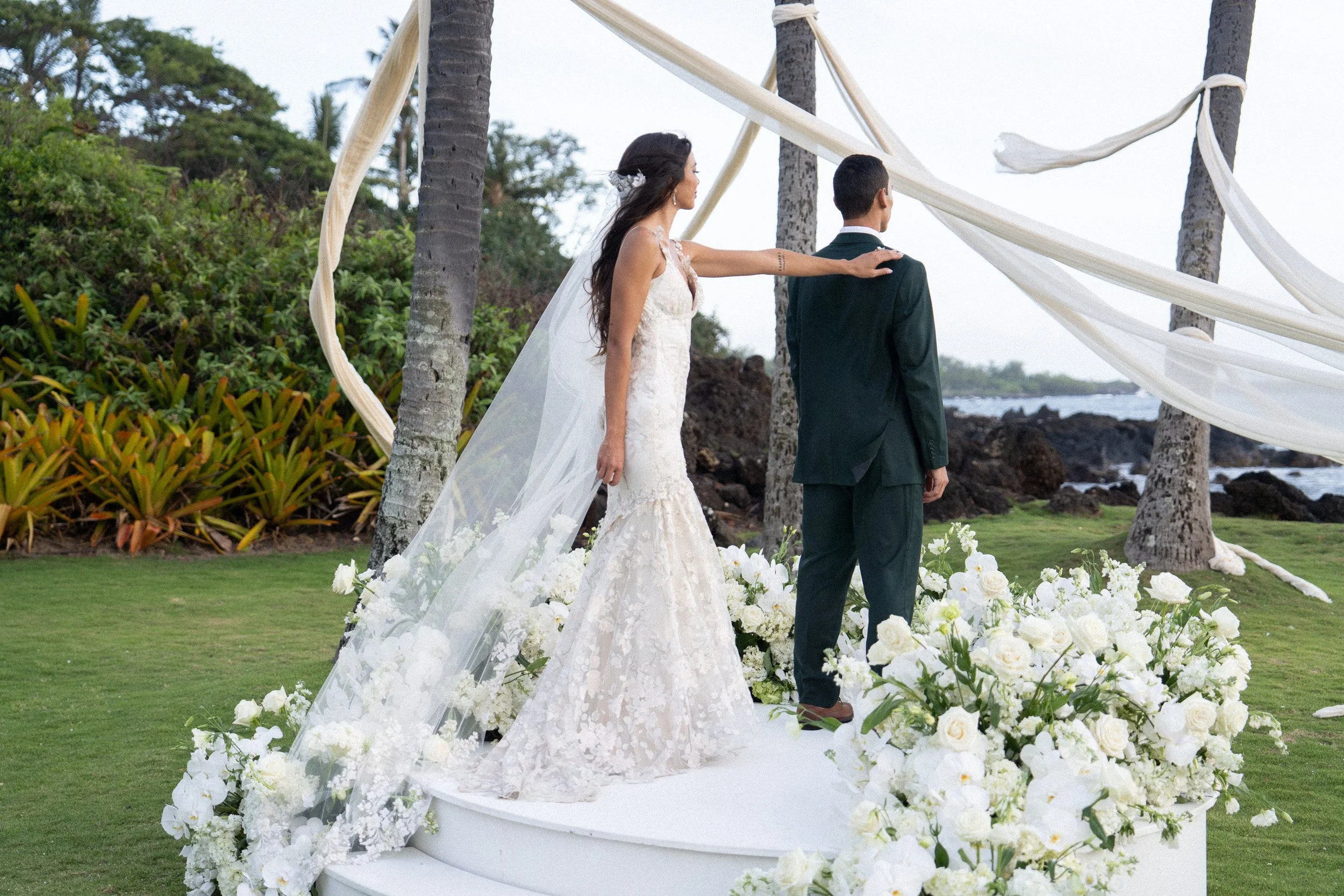 A bride and groom stand under a decorated wedding arch made of palm trees and flowing fabric, surrounded by white flowers on a grassy outdoor setting near the coast.