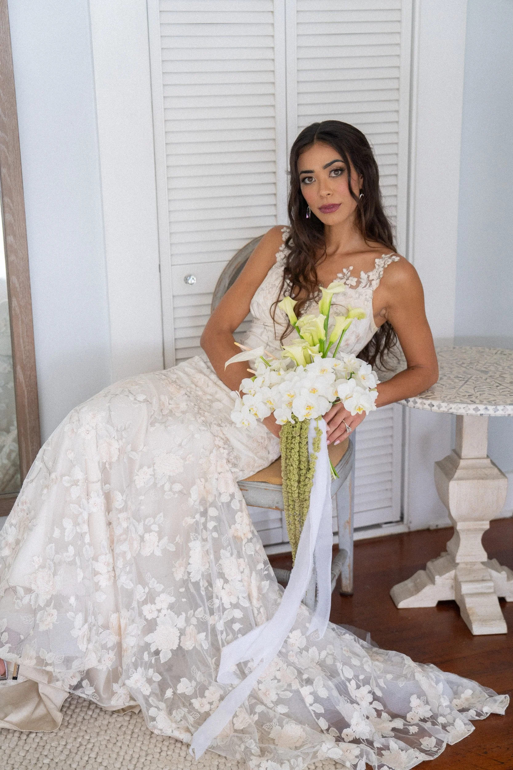 A woman in a white lace wedding dress with floral pattern holds a bouquet of calla lilies and white orchids, sitting on a chair in a room with white louvered doors and a small table.
