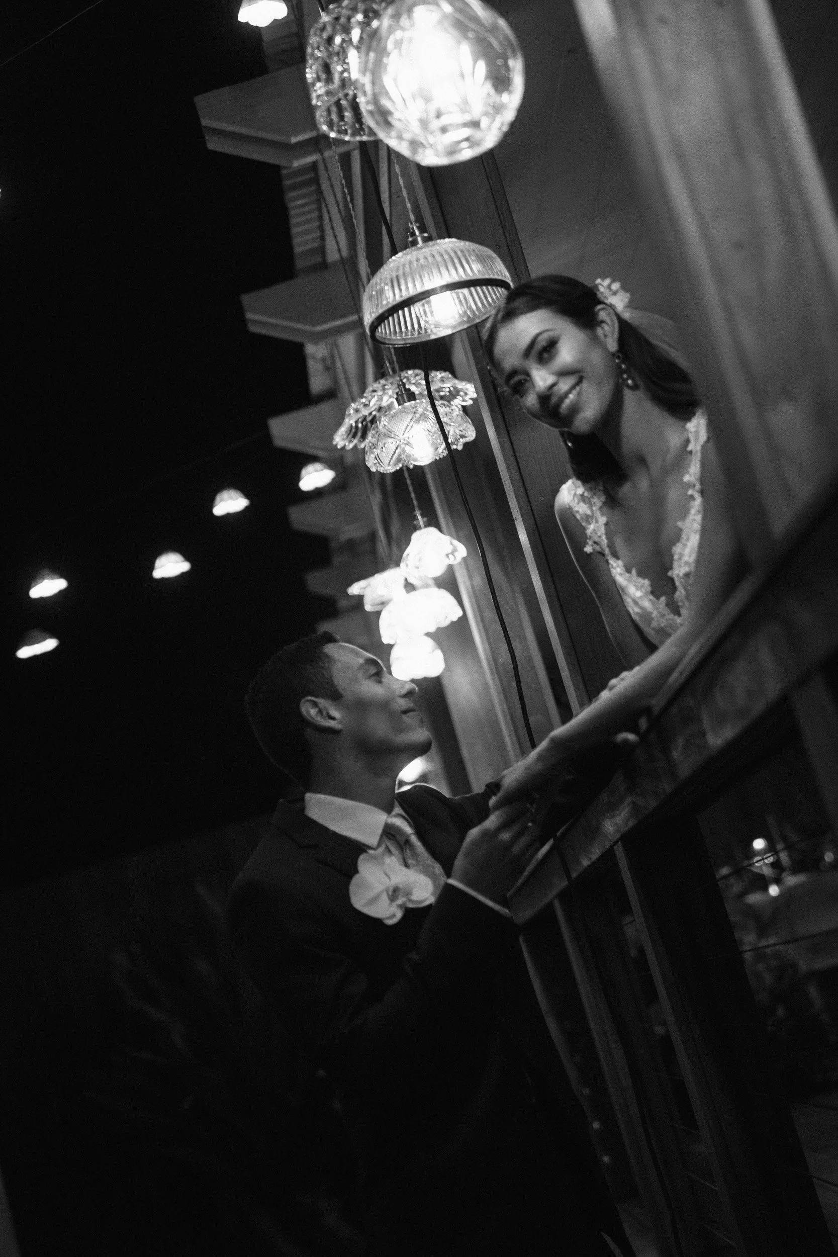 A black and white photo of a smiling woman with dark hair and earrings, talking to a man dressed in a suit, both sitting at a table in a dimly lit restaurant with pendant lamps hanging above.