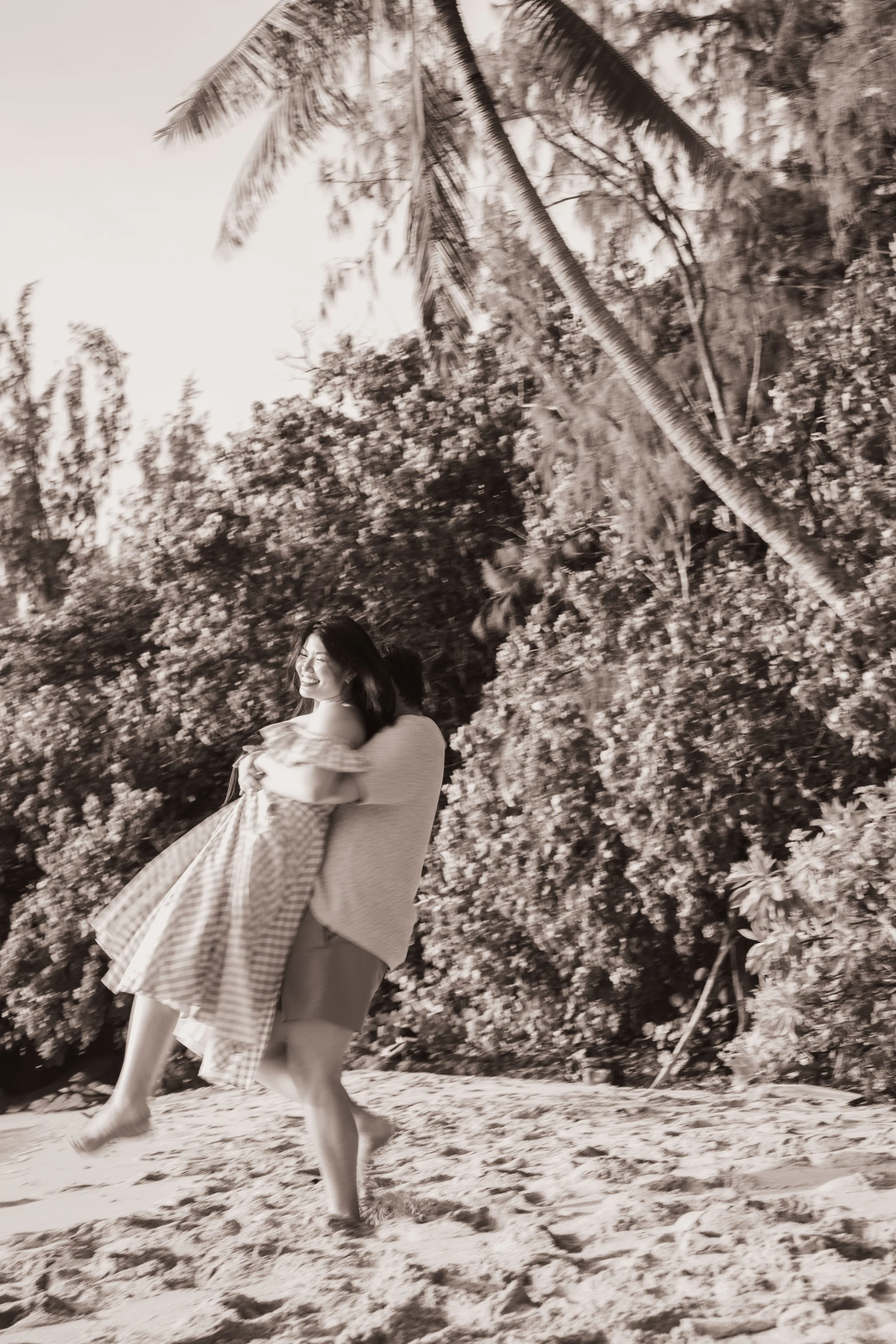 A woman holding a small girl on a sandy beach with lush trees and a palm tree in the background, all in black and white.