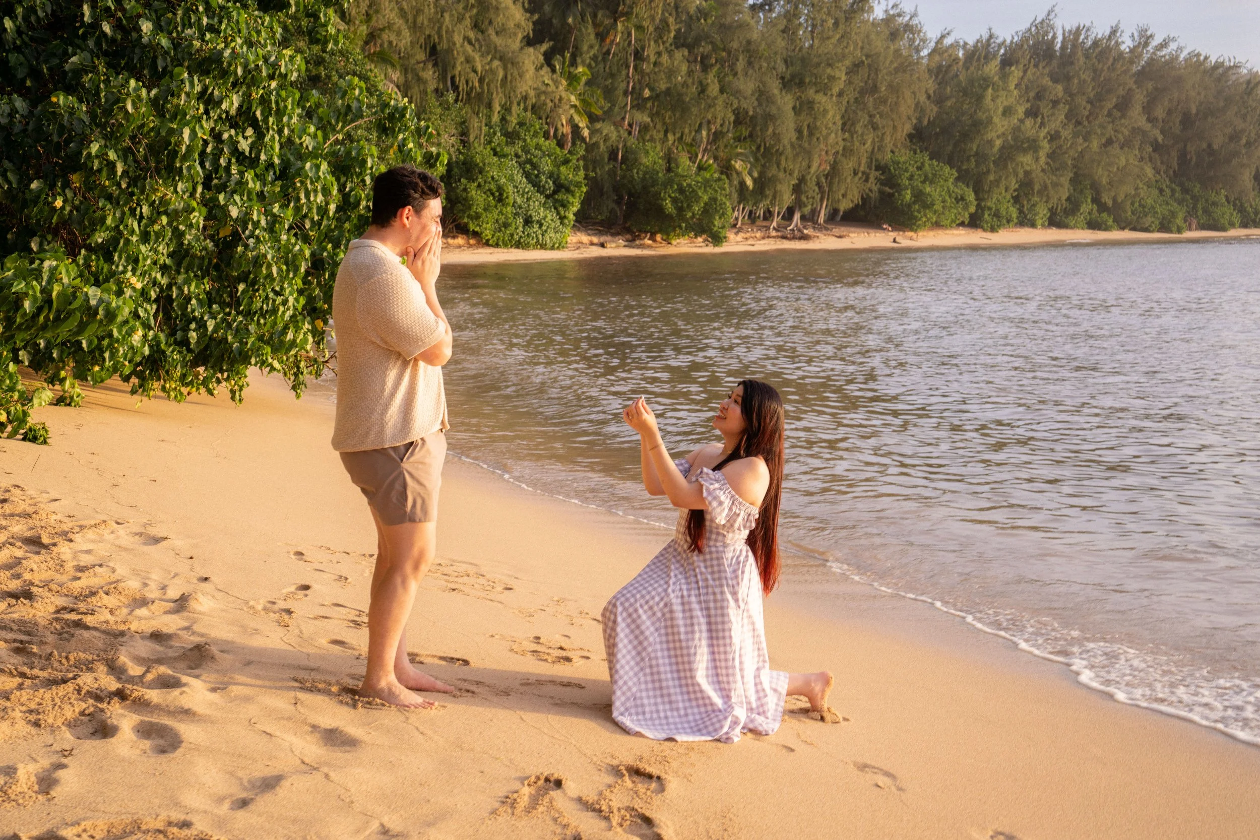 A woman is proposing marriage to a man on a beach at Kawela , with the woman kneeling and holding out her hand, while the man covers his mouth in surprise or emotion. The beach is sandy with green trees and water in the background during the daytime.