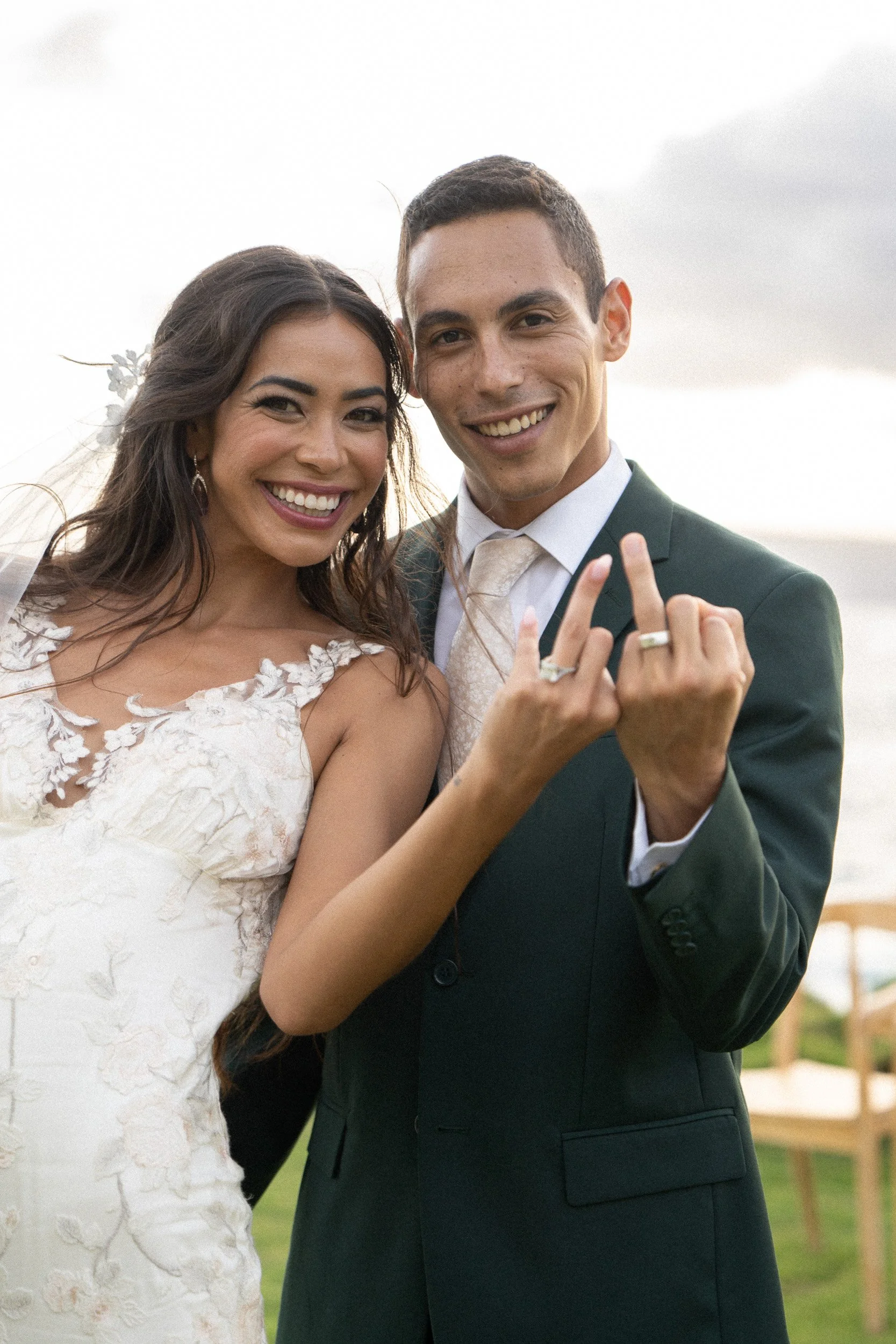 A happy bride and groom smiling and showing their wedding rings outdoors, with a cloudy sky in the background.