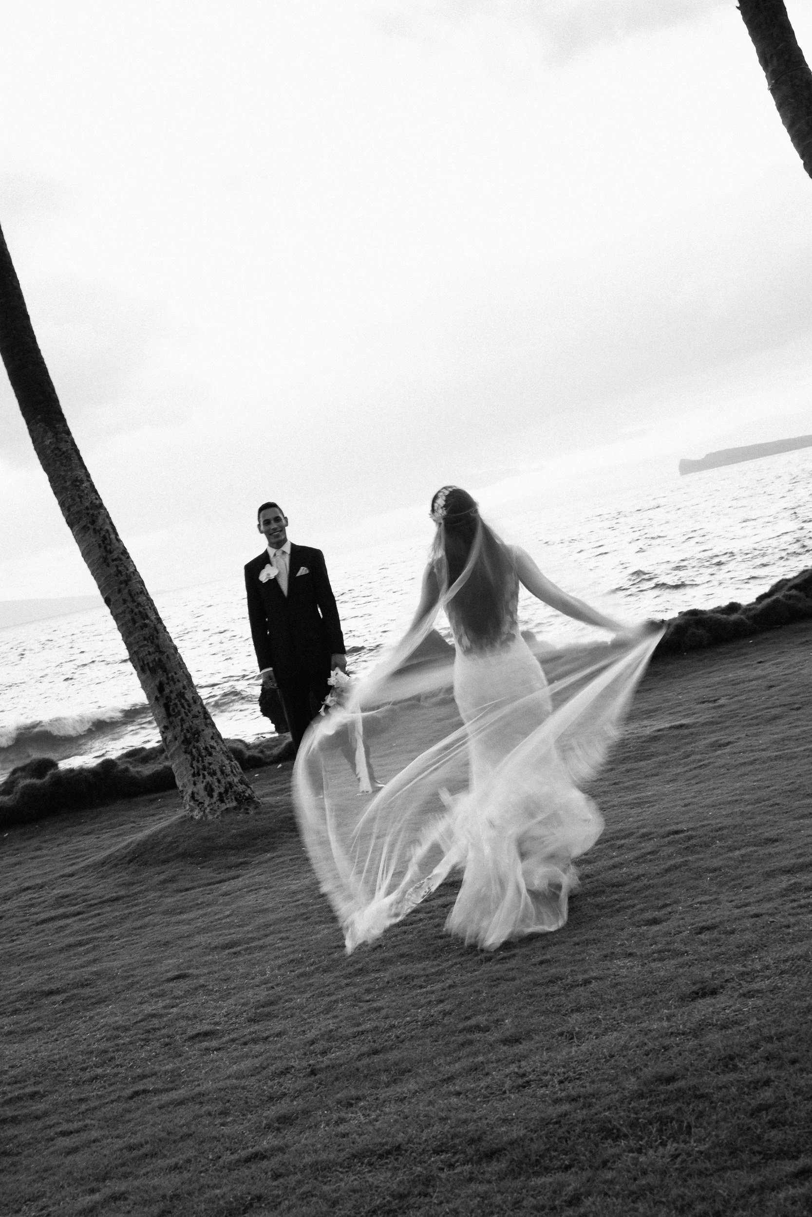 A black and white photo of a bride in a flowing dress and veil dancing on a beach near the ocean, with a groom in a suit and tie holding a bouquet, surrounded by palm trees.