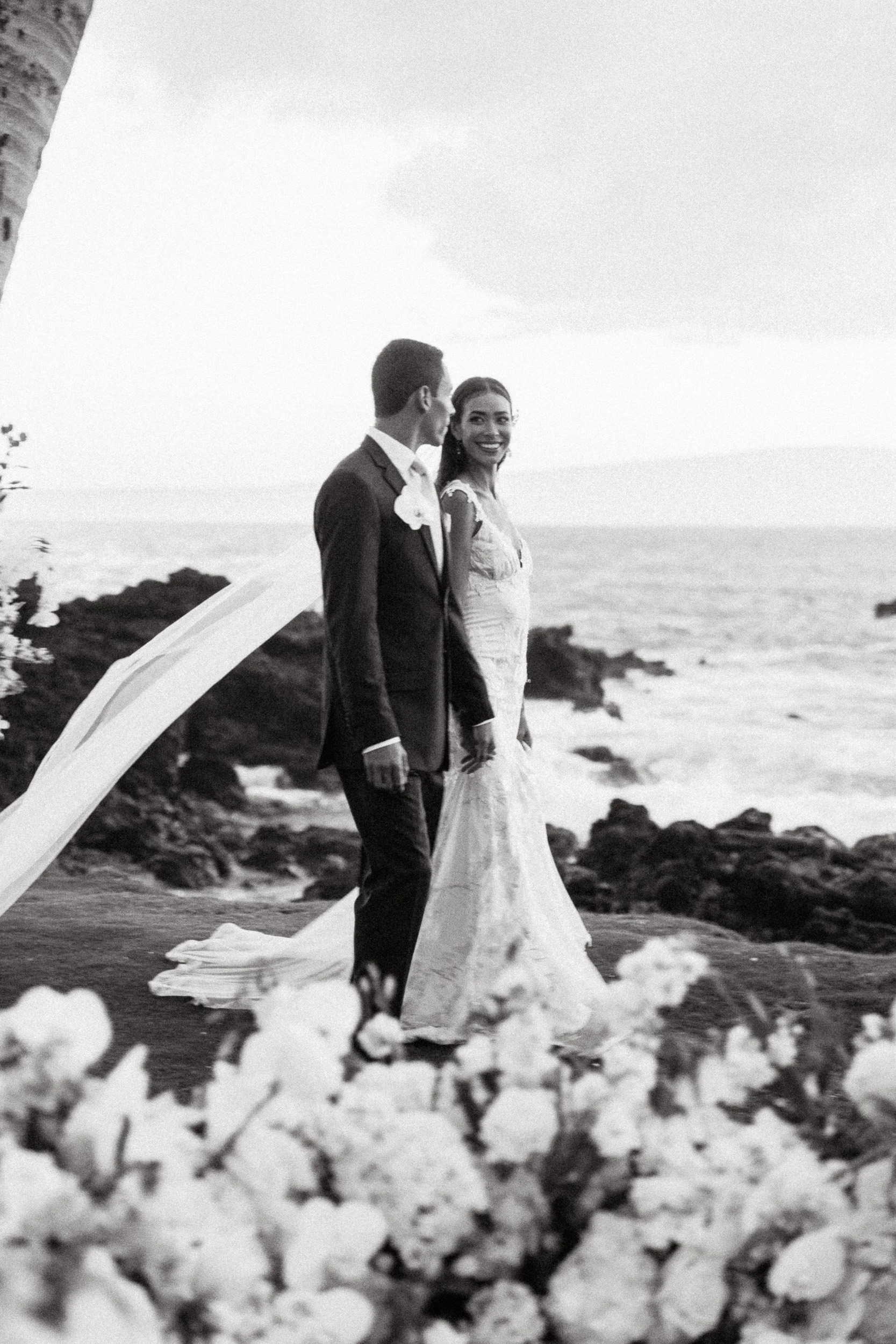 A wedding couple walking along a rocky shoreline with the ocean in the background, black and white photo.