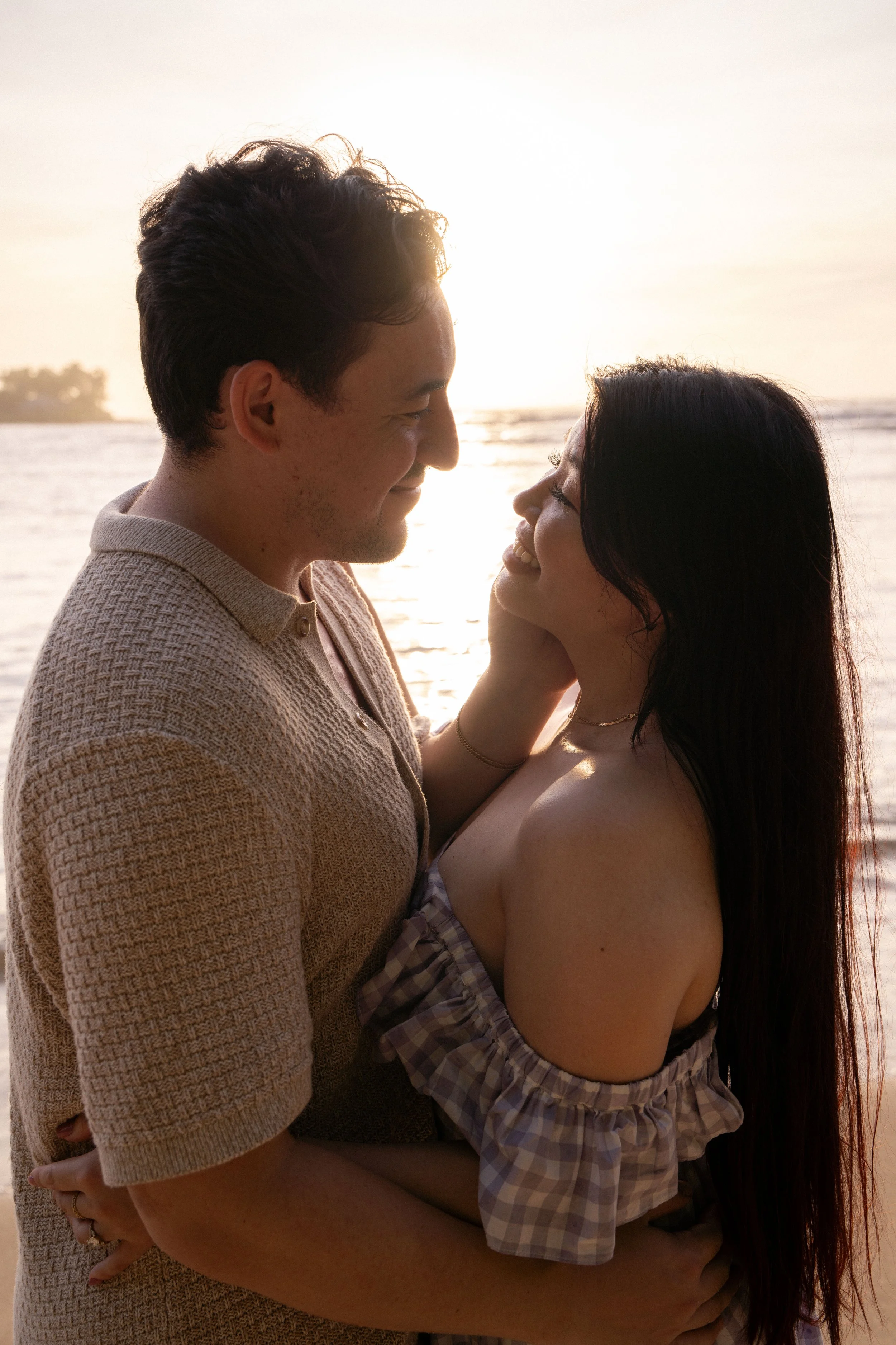 A man and woman standing close together by the water during sunset, smiling at each other.