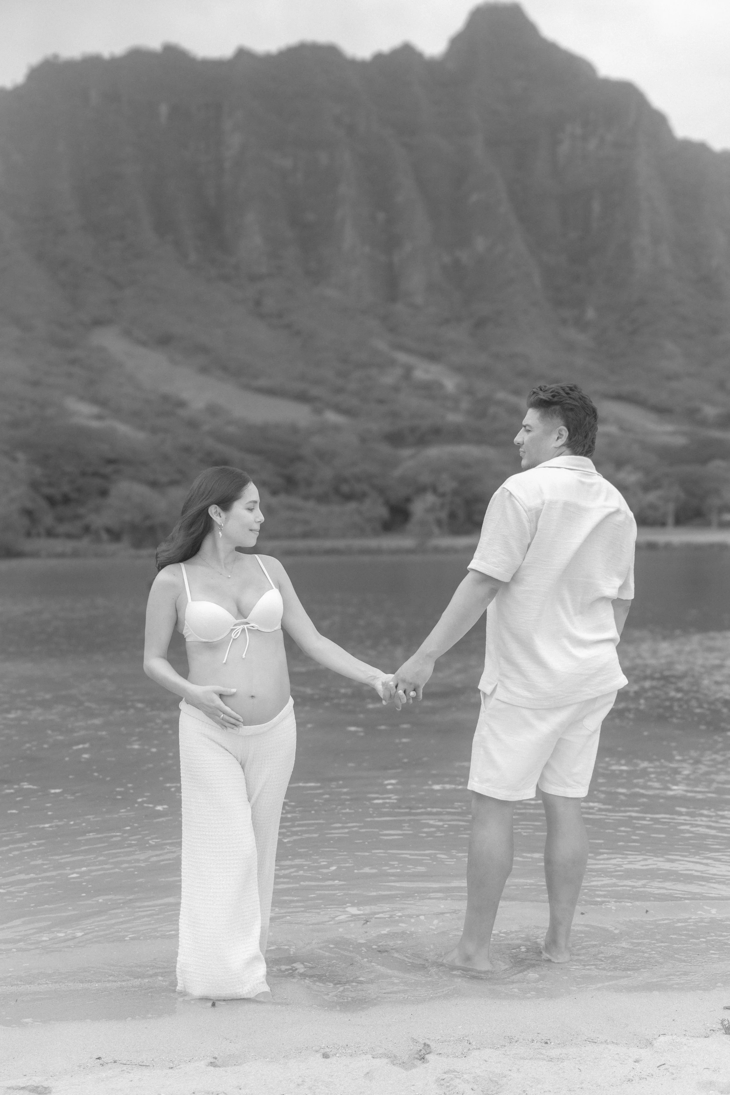 A black and white photo of a couple holding hands on a beach. The woman is wearing a white bikini top and a long skirt, while the man is dressed in a light shirt and shorts. They are facing each other with a large mountain in the background.