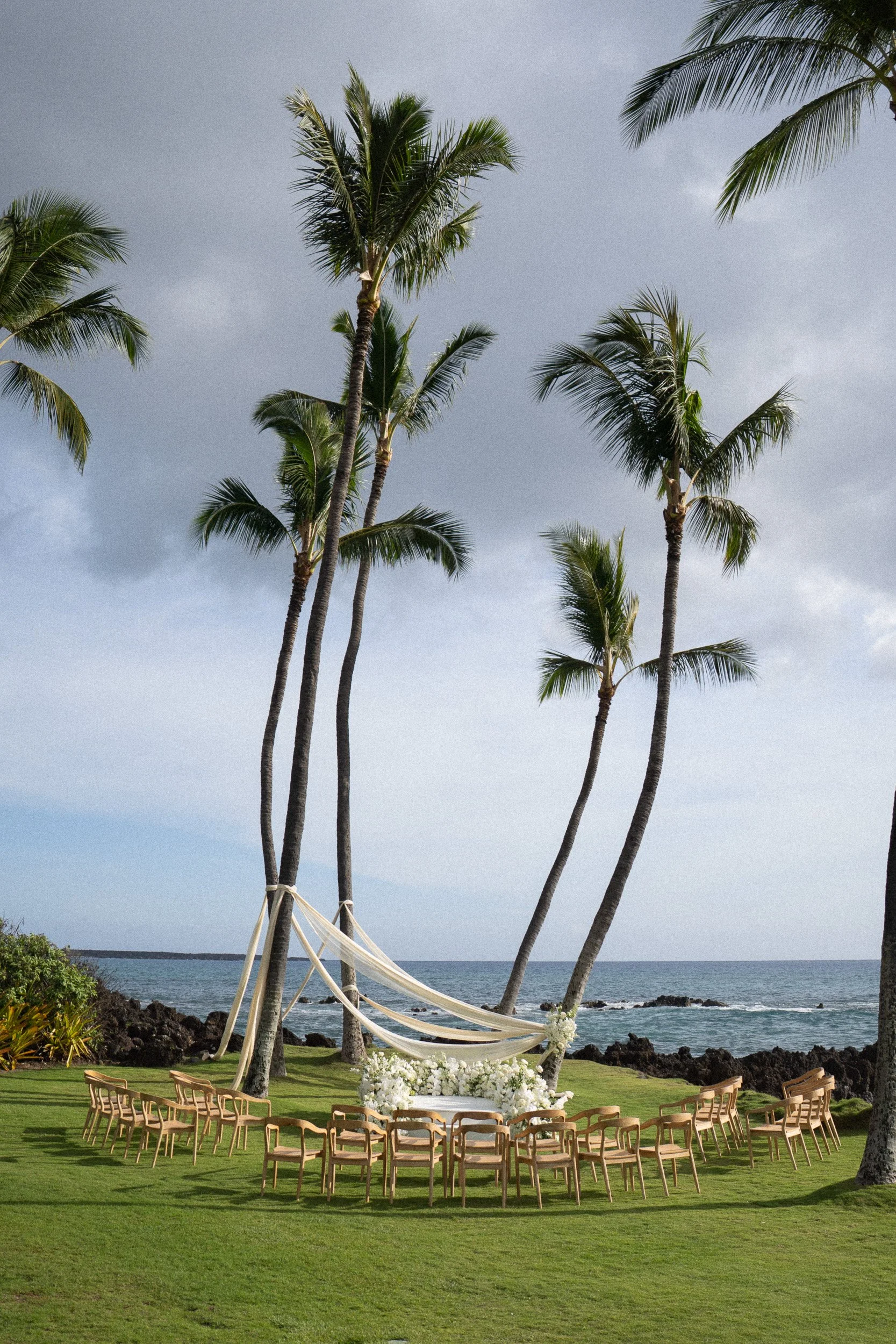 Outdoor wedding ceremony setup on green grass with chairs arranged in rows, decorated with white flowers and a white canopy, palm trees in the background near the ocean, overcast sky