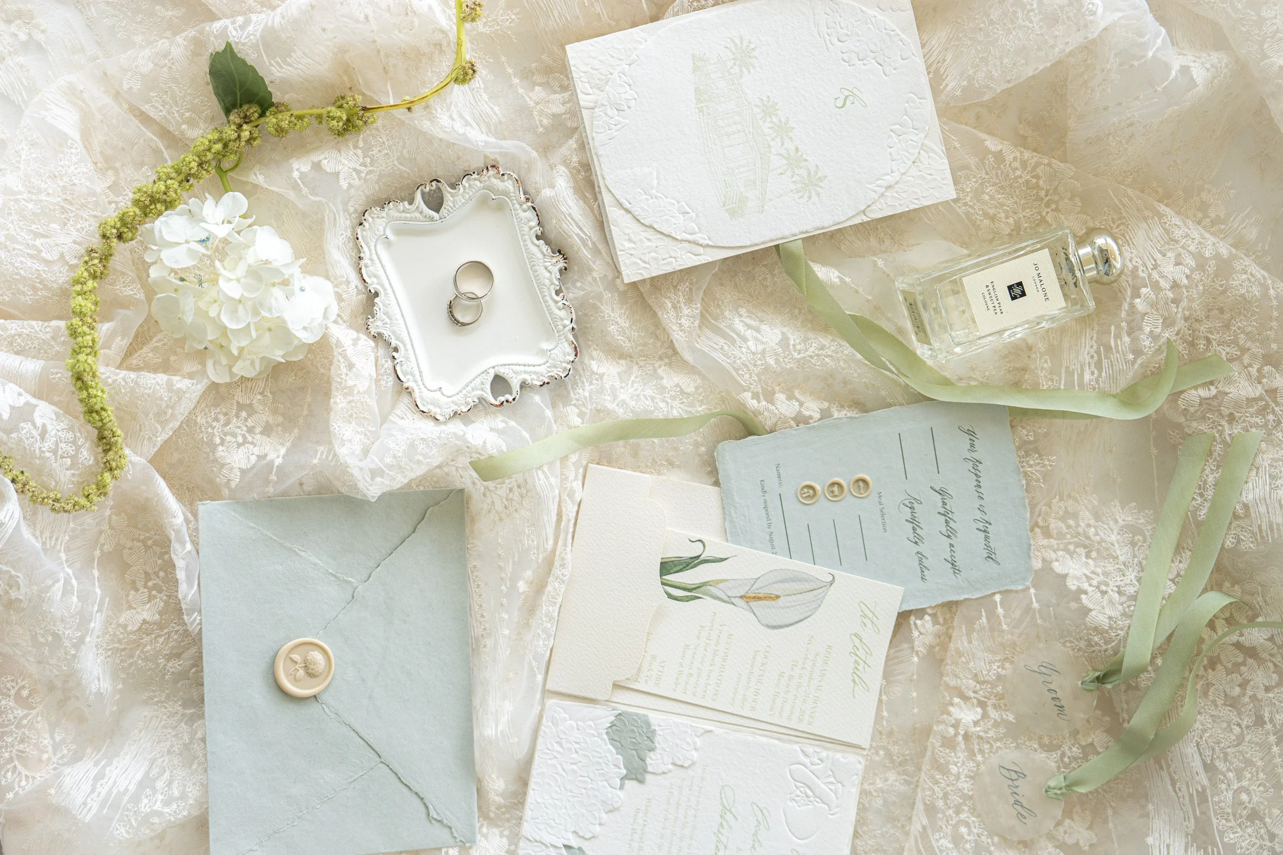 A flat lay of wedding stationary and accessories, including wedding invitations, rings, a small dish, a bottle of perfume, buttons, and some flowers on lace fabric.