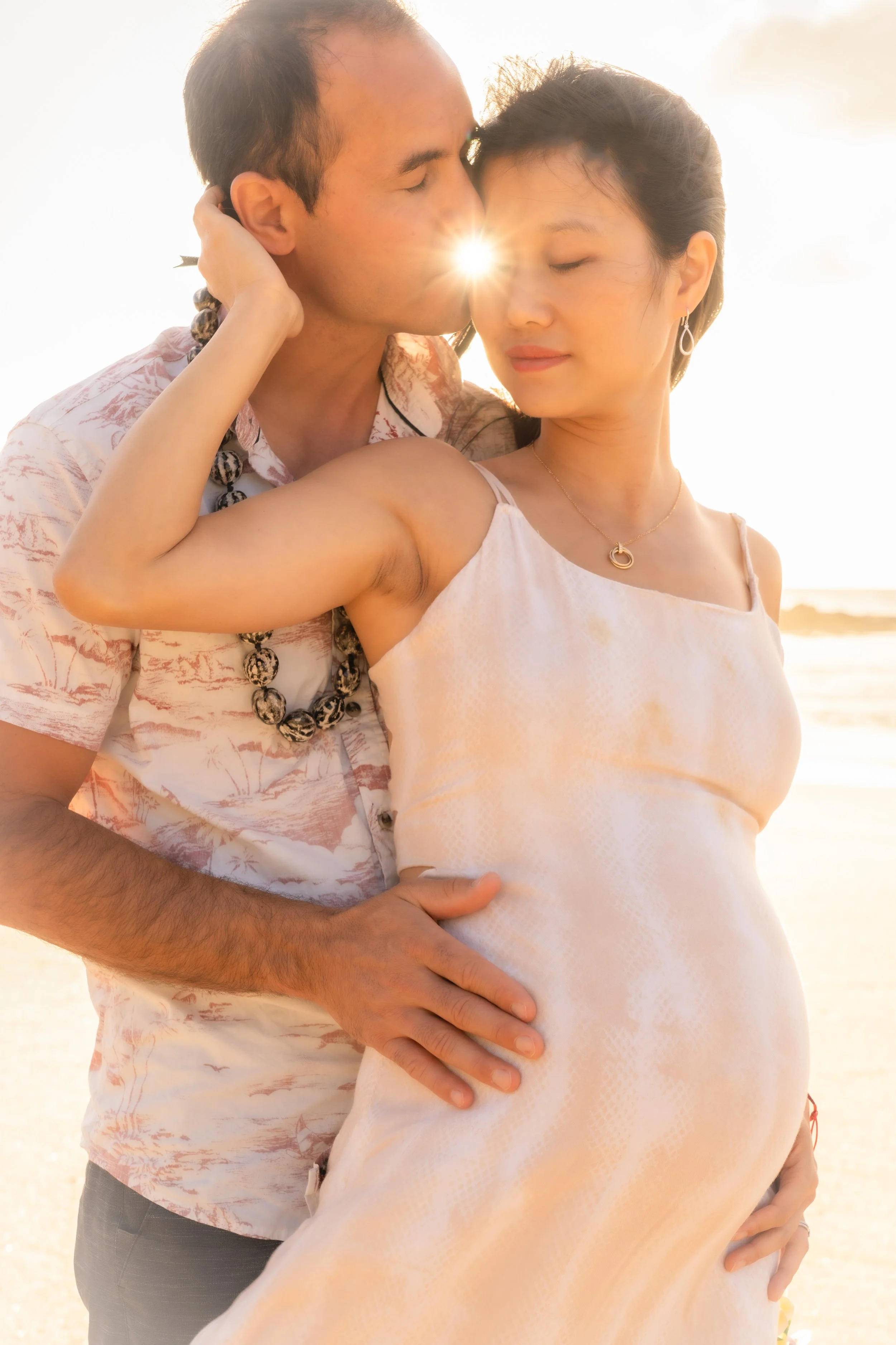 A couple on the beach during sunset at keiki beach, with the man kissing the woman's forehead while she gently holds her pregnant belly.