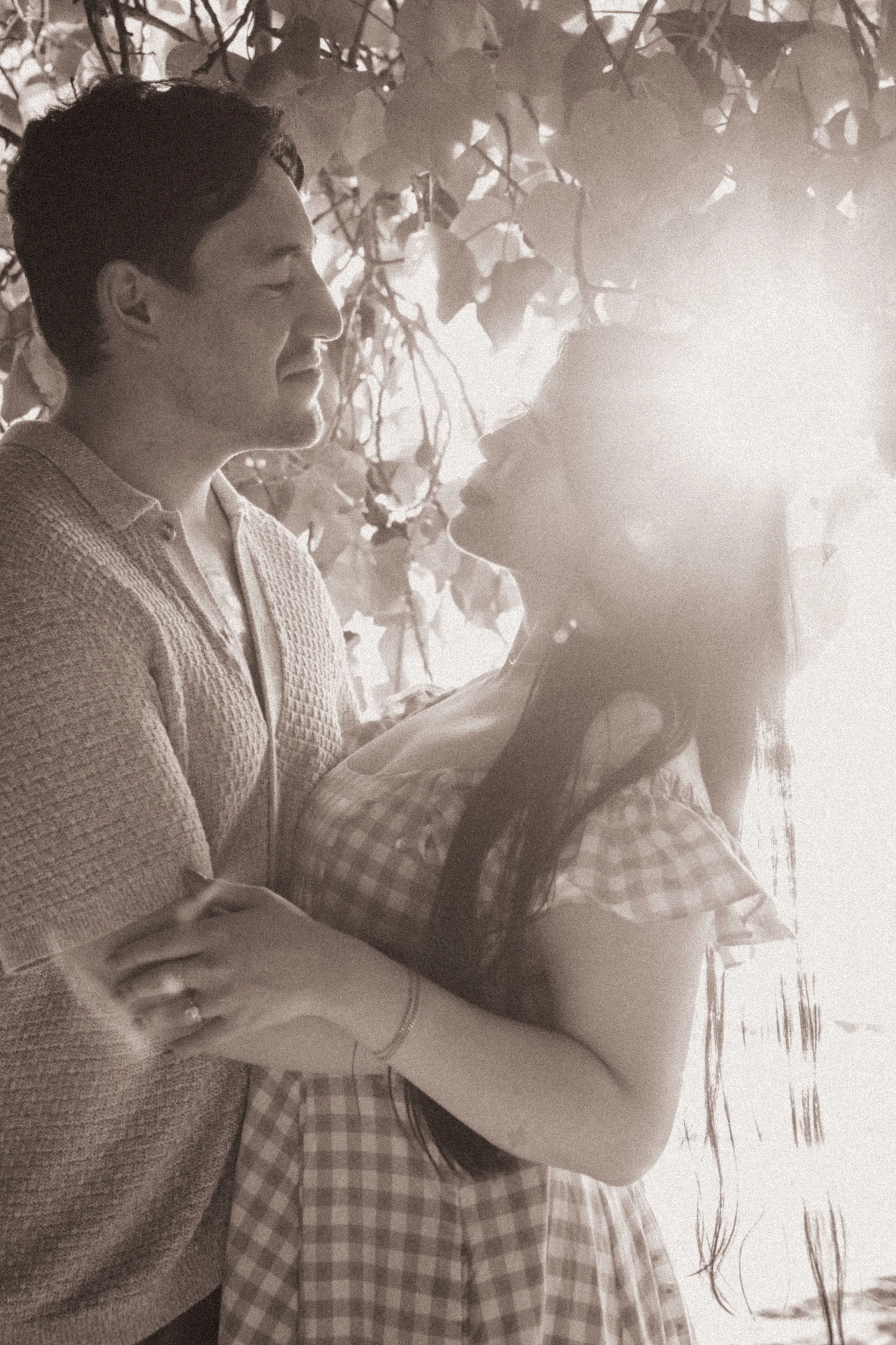 A black-and-white photo of a couple sharing a close moment outdoors, with the man smiling and the woman wearing glasses, surrounded by trees and plants.
