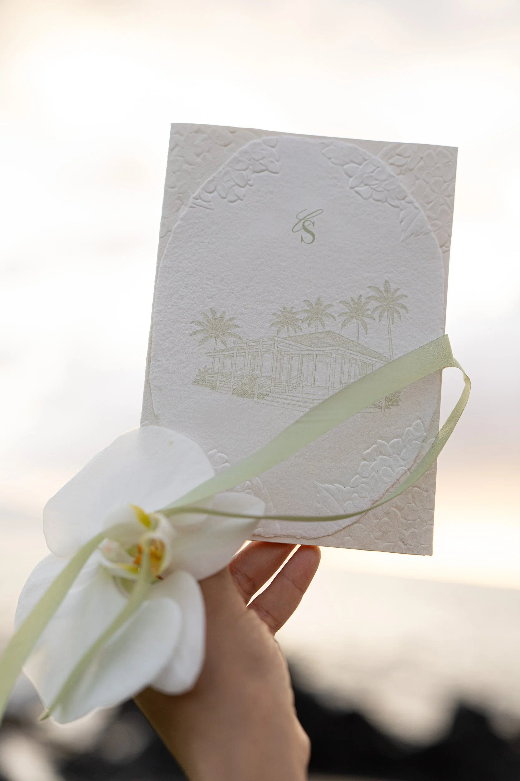 A hand holding a wedding invitation card with a white orchid flower in front, and a light, blurry background.