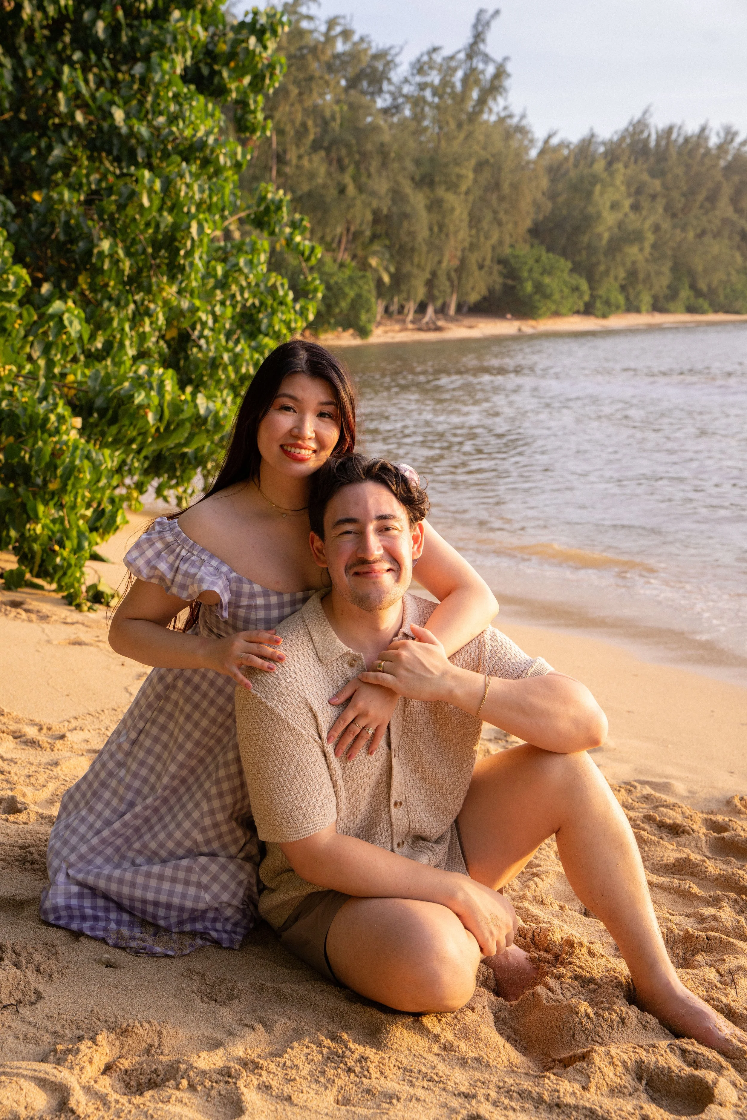 Two friends sitting on a sandy beach, smiling, near the water with trees in the background during sunset.