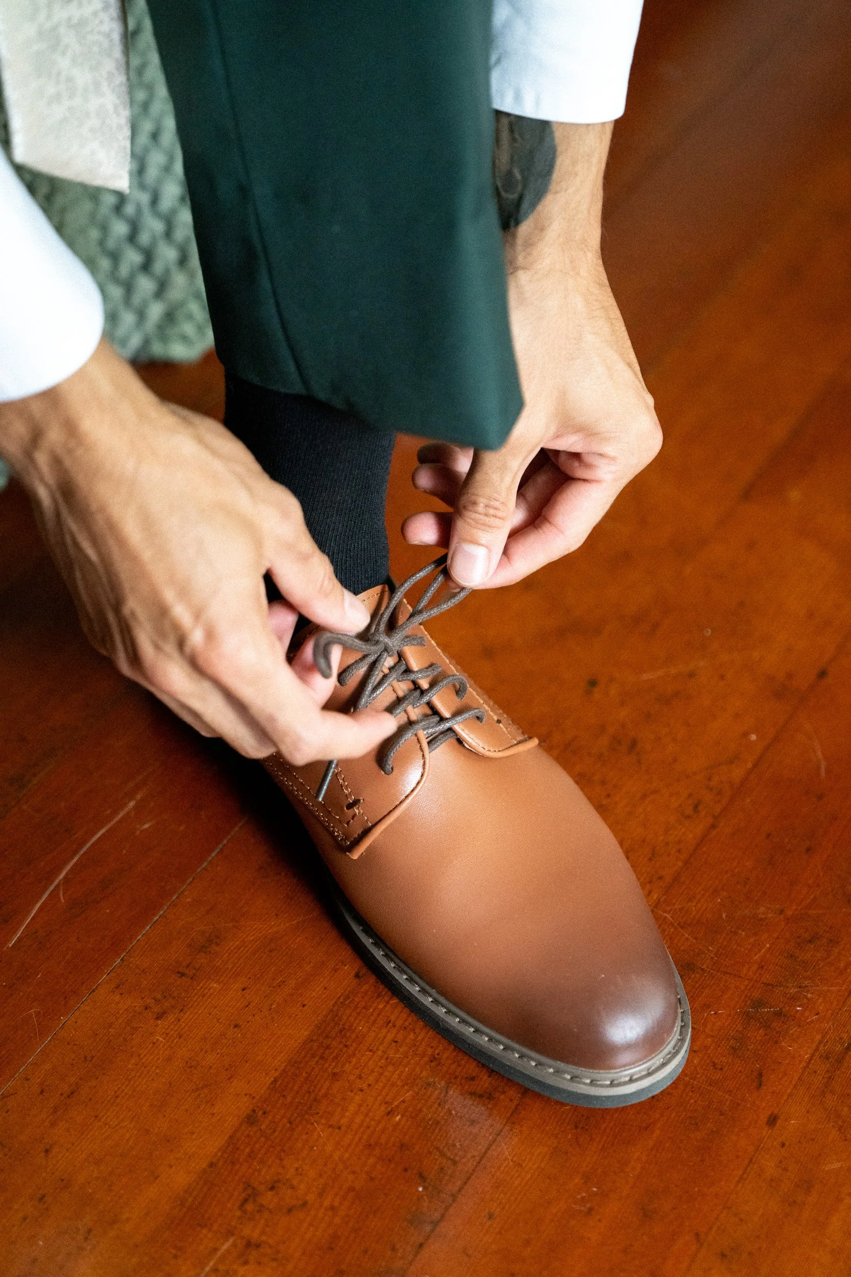 A person is tying the laces of a tan leather dress shoe while sitting on a wooden floor. The person is wearing black socks, dark green or black pants, and a white shirt. The shoe is well-polished and new, and the person's hands are focused on lacing 