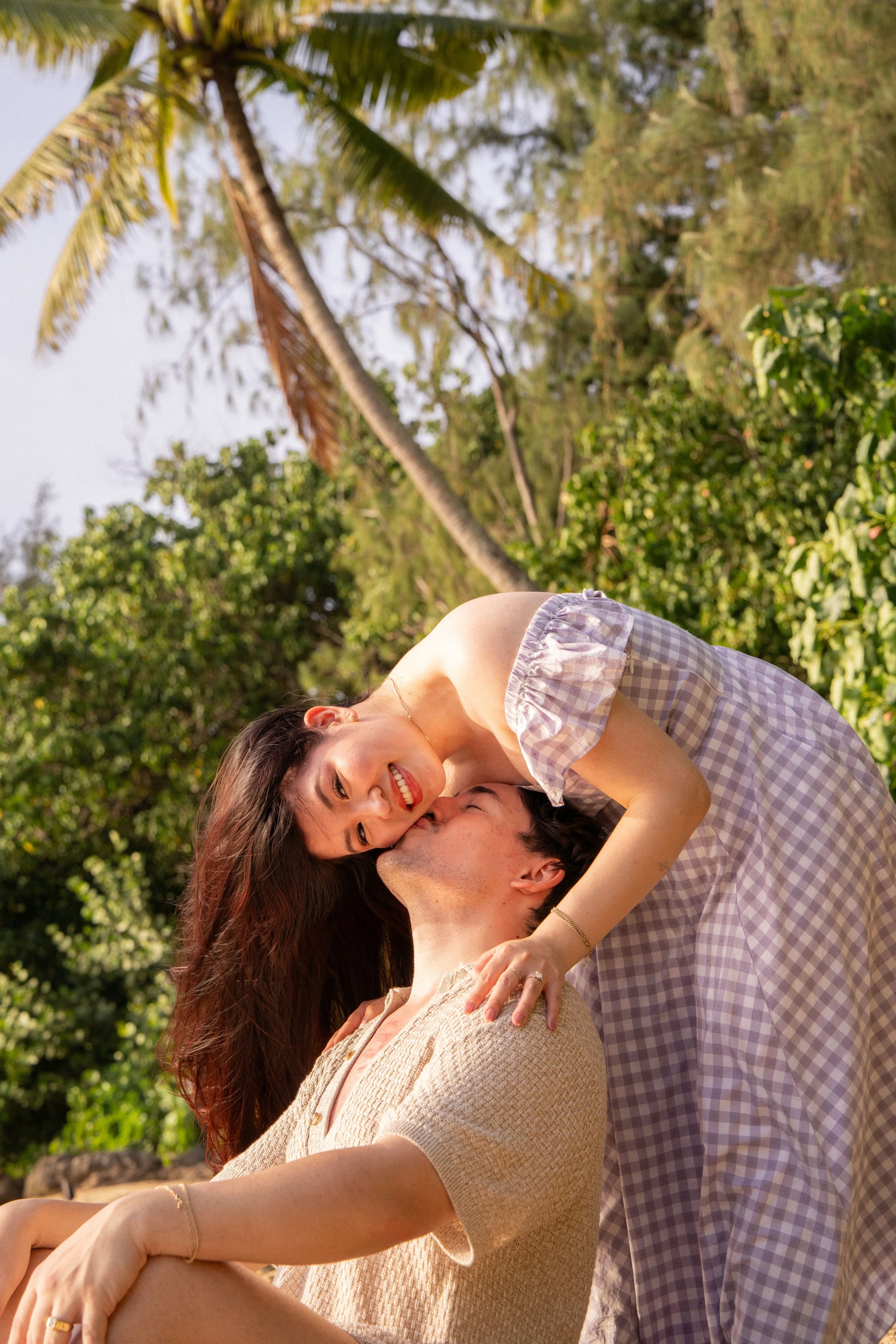 A couple sharing a romantic moment outdoors with lush green trees and a palm tree in the background. She is sitting on his lap, smiling, with her arms around his neck. He is looking up at her, smiling.