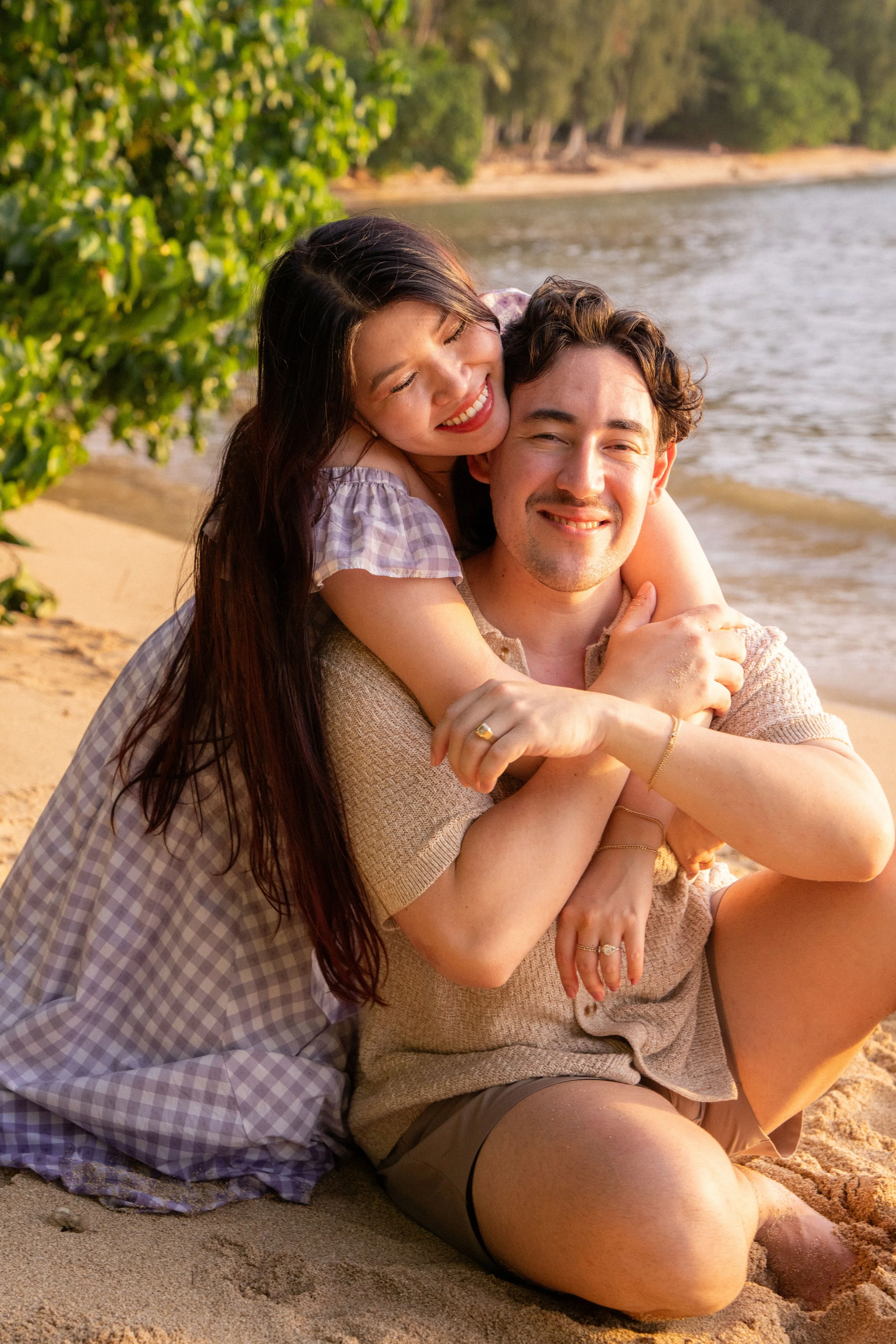 A happy couple sitting on the beach, enjoying each other's company during sunset. The woman is on the man's back, hugging him, and both are smiling. The woman has long dark hair and is wearing a checkered dress, while the man has curly hair and is we
