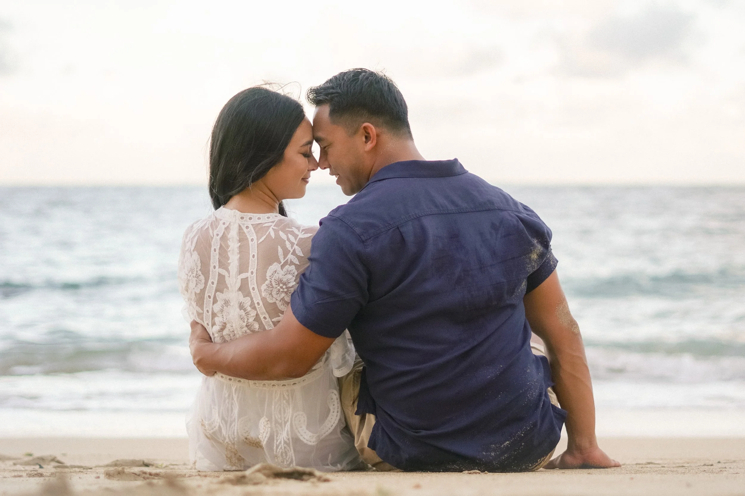 A couple sitting on the beach, facing each other with foreheads touching, smiling, and holding hands, during a cloudy day by the ocean. candid couple portrait in Haleiwa with ocean view.
