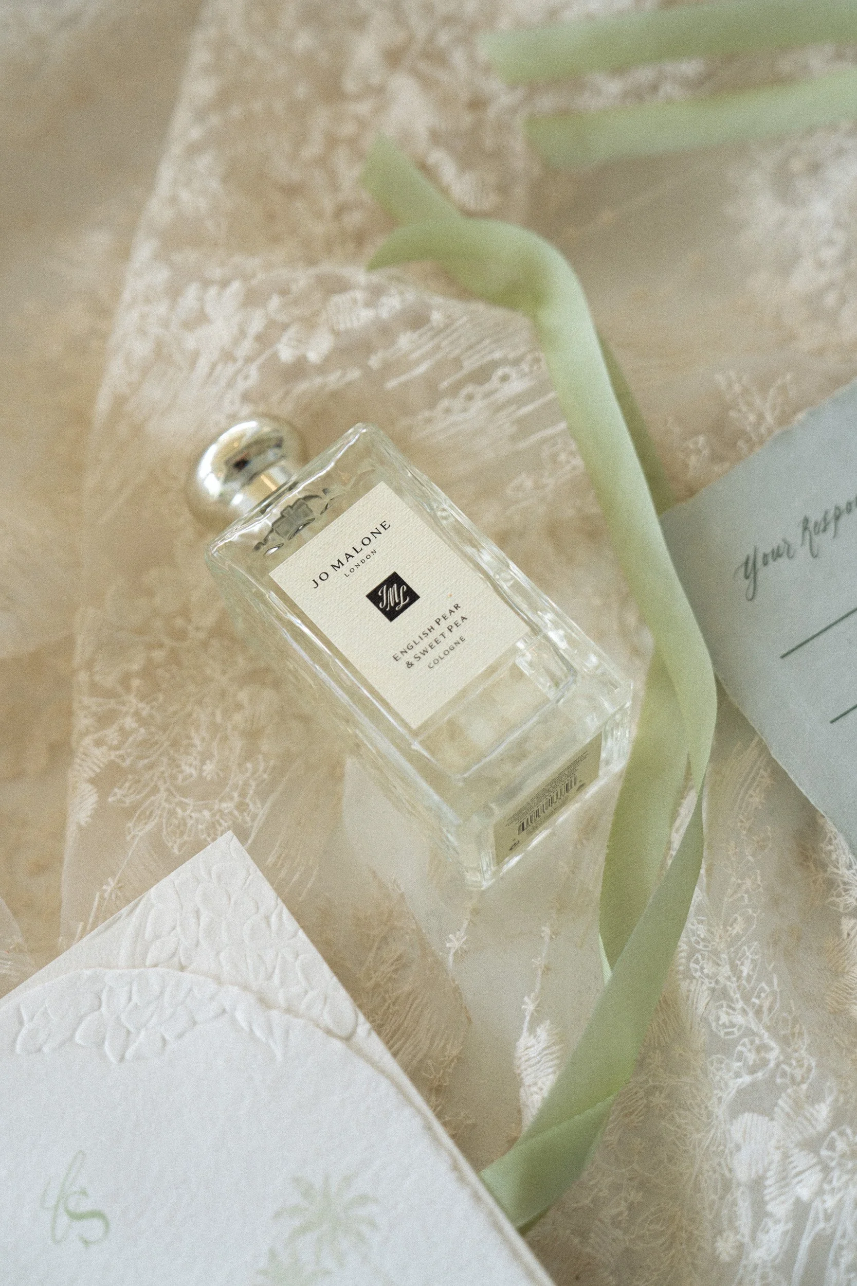 A clear Jo Malone perfume bottle, a green tulip, and a greeting card on a lace tablecloth.