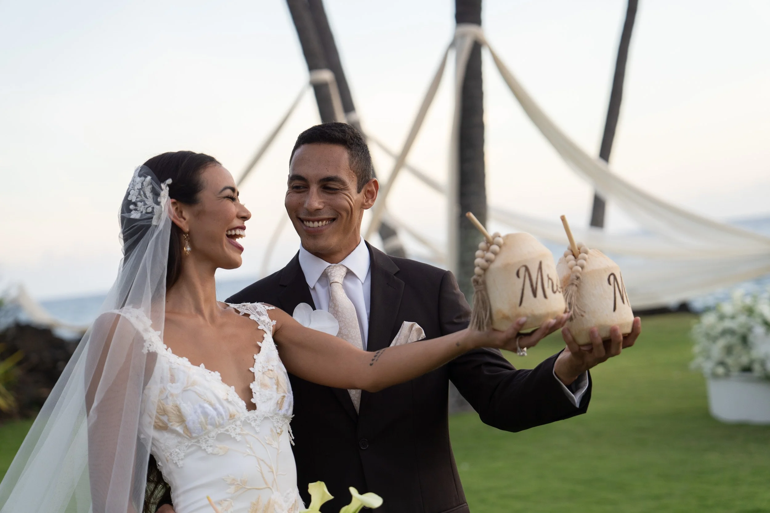 A bride and groom smiling and holding coconuts with straws during their outdoor wedding celebration.