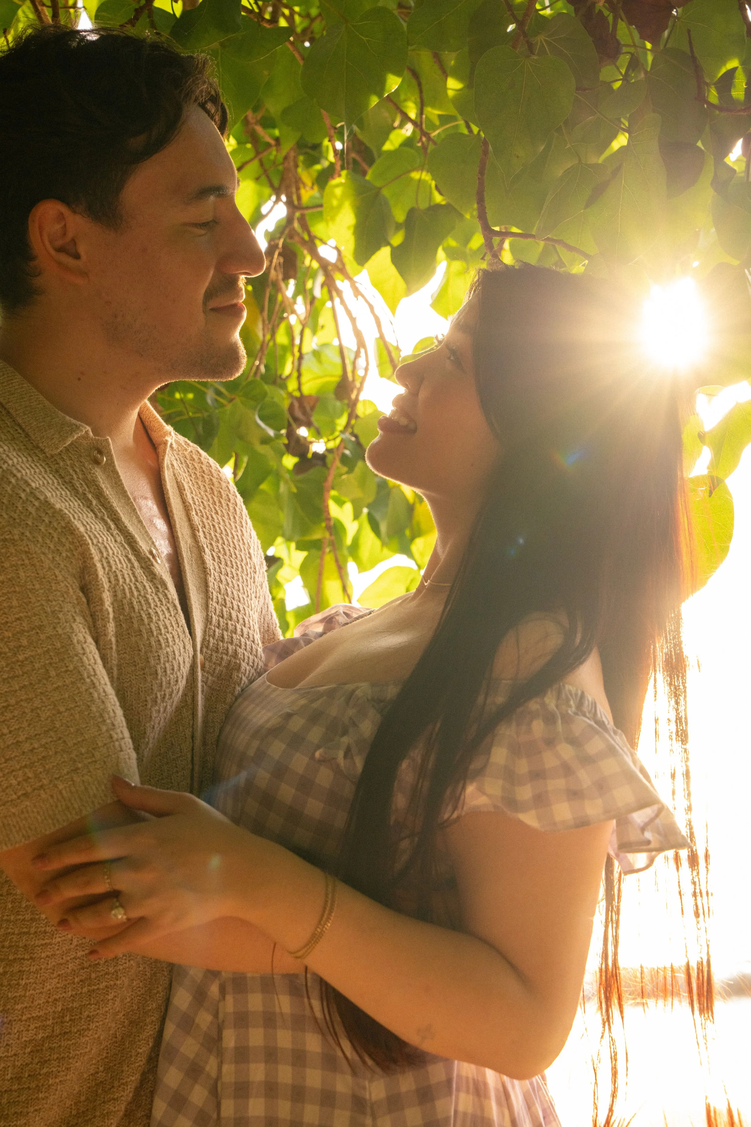 A man and woman looking at each other affectionately outdoors, with sunlight shining through leaves in the background.