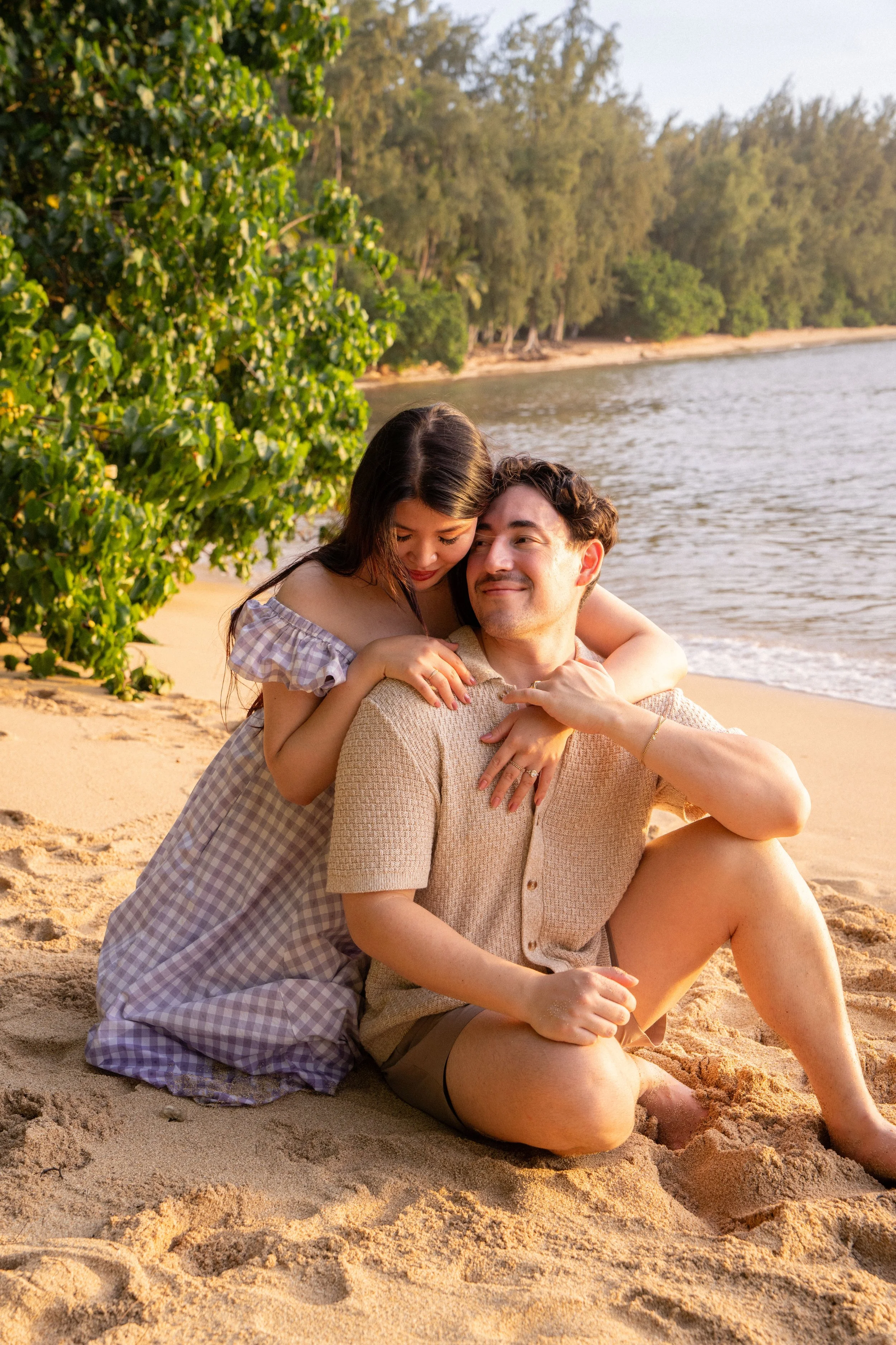 A couple sitting on sandy beach near water, woman hugging man from behind, trees in background, warm sunset lighting.