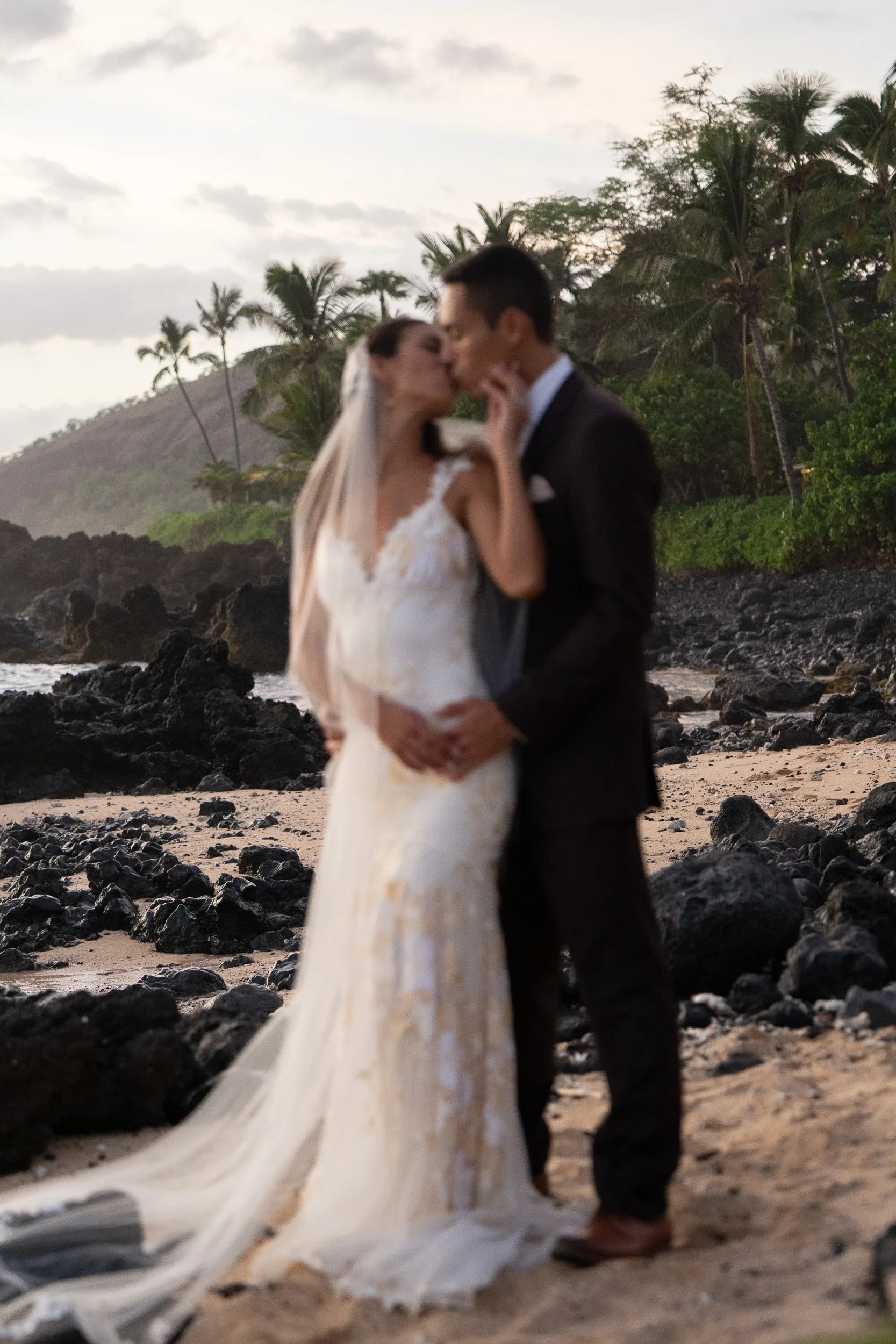 A bride and groom kissing on a rocky beach with palm trees in the background during sunset.