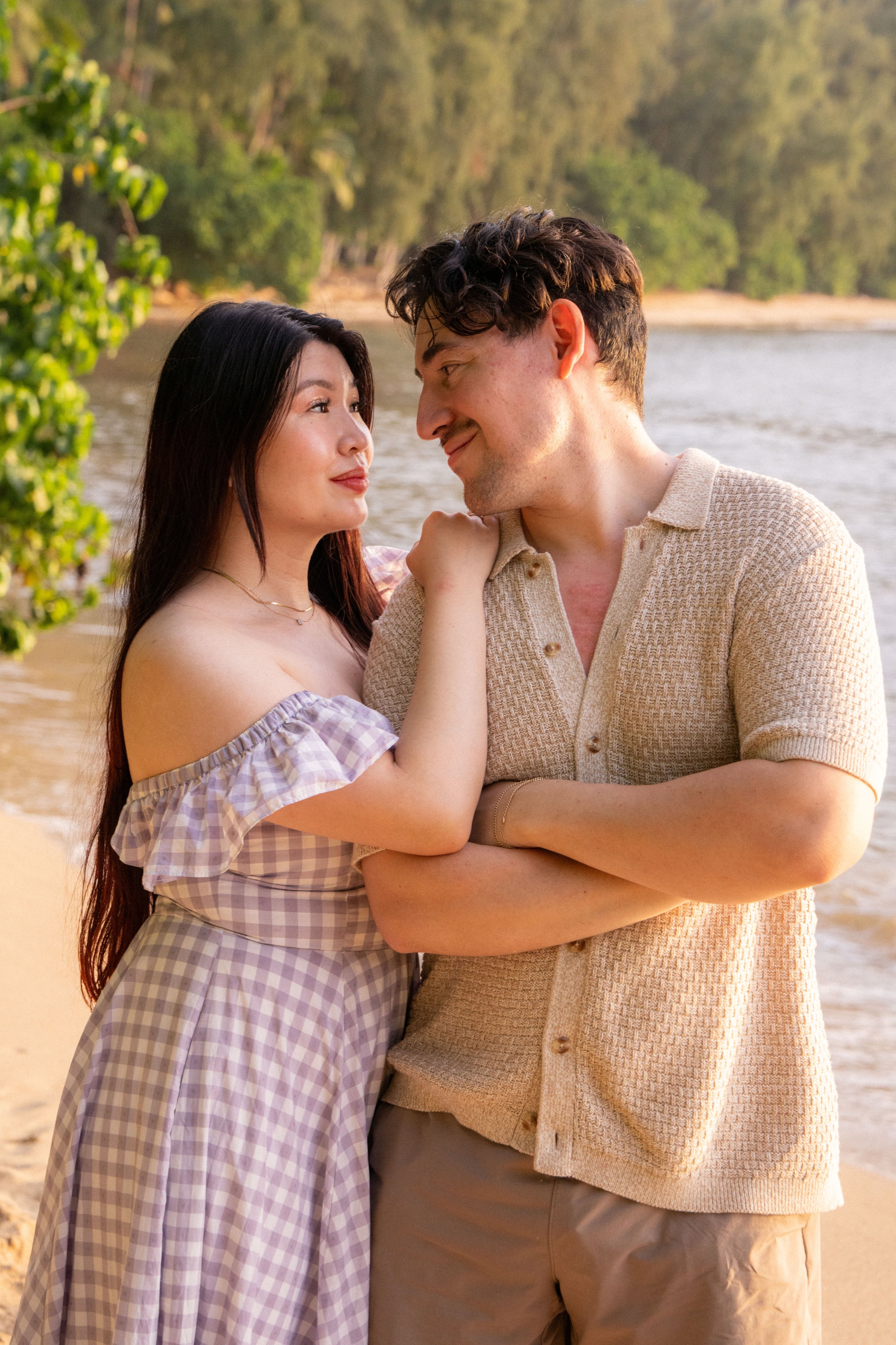A couple standing close together near a body of water, gazing into each other's eyes with smiles. The woman has long dark hair, wearing a gingham off-shoulder dress. The man has short curly hair, wearing a beige textured shirt. They are outdoors with