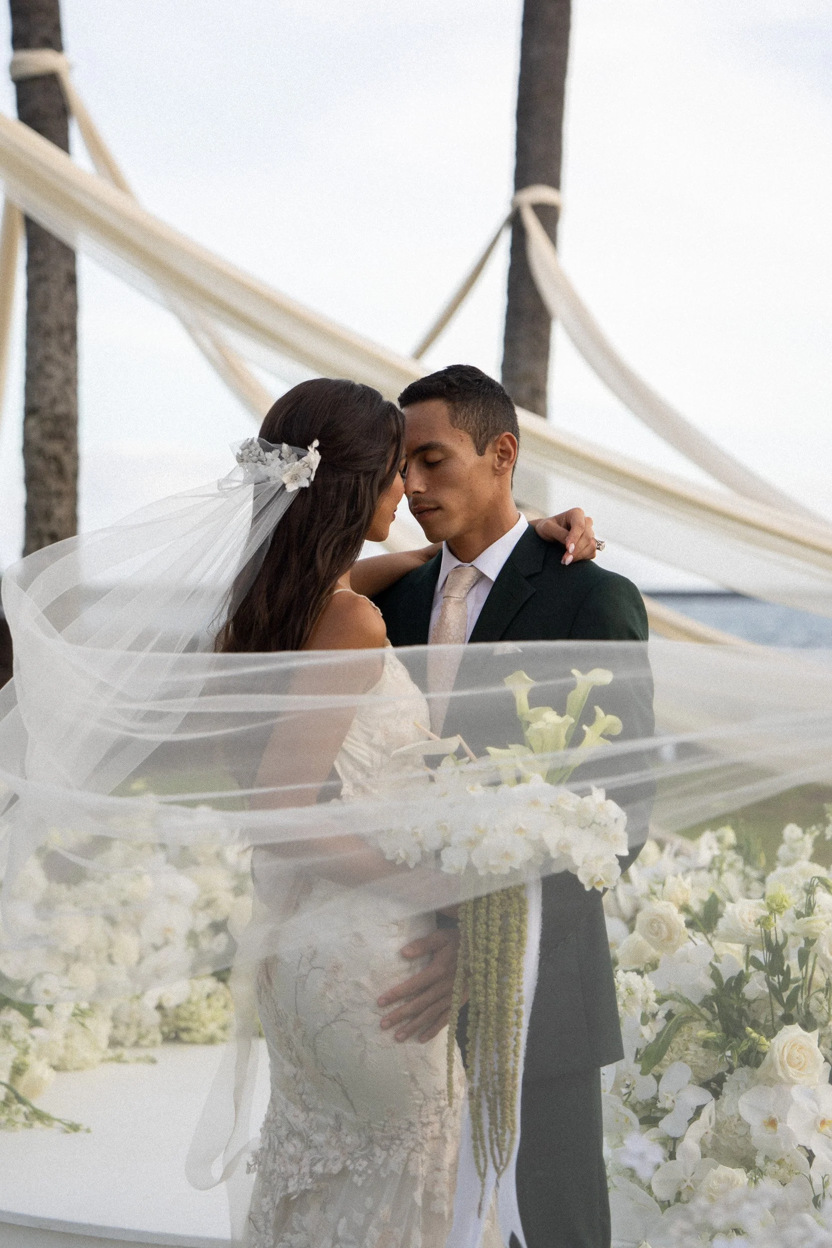 A bride and groom are standing close together during their wedding ceremony, with their foreheads touching. The bride is wearing a white lace gown and a veil with flowers, while the groom is dressed in a black suit, white shirt, and a light-colored t