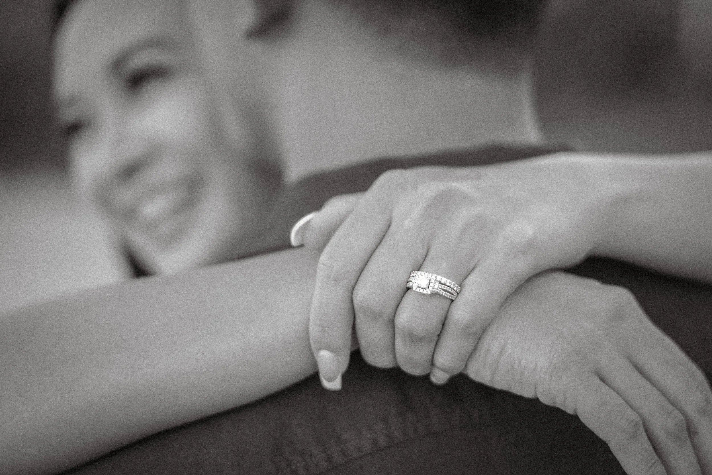 A woman smiling and showing her engagement ring on her finger while resting her arms on her partner's arm.