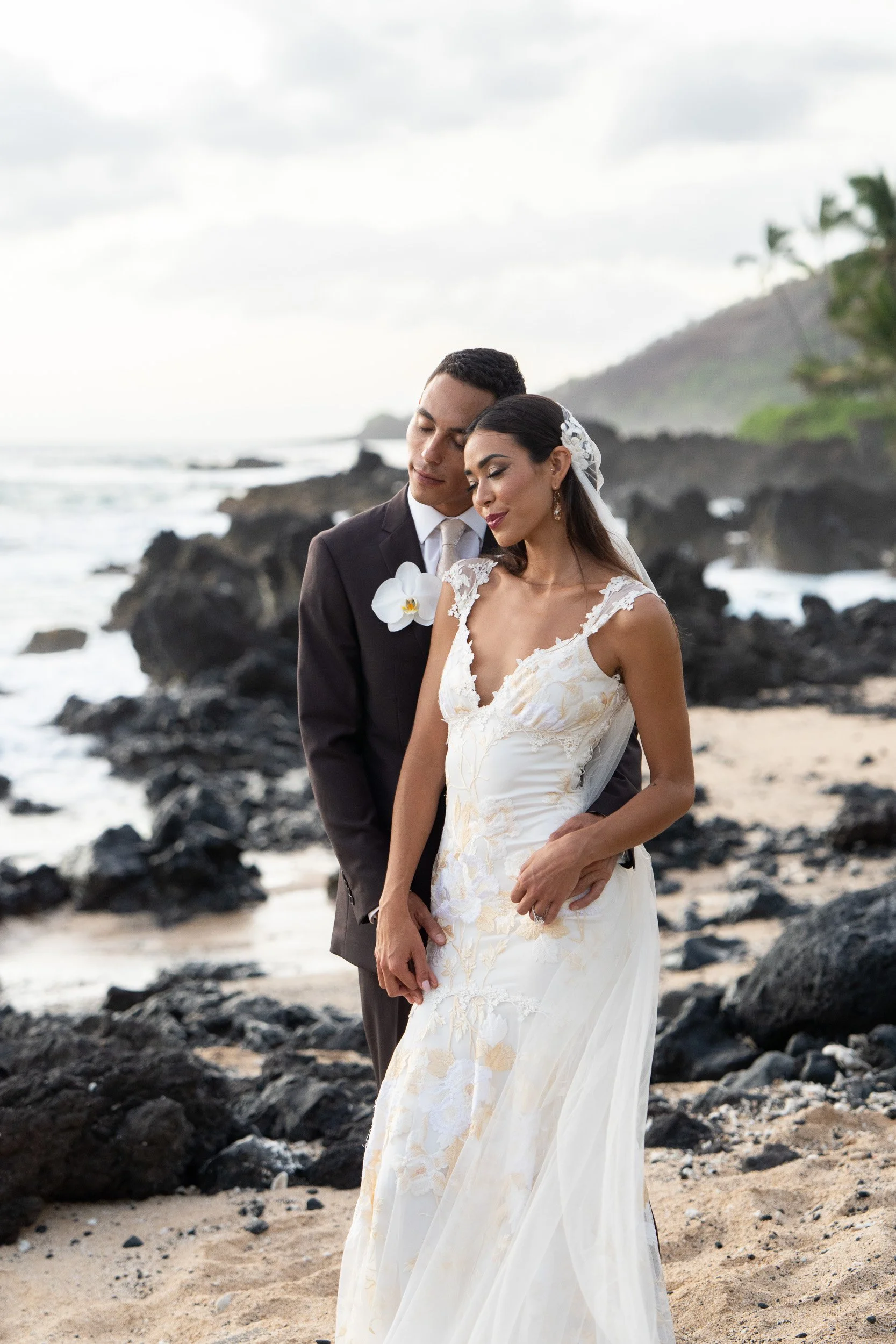 A newlywed couple in wedding attire standing on a rocky beach with ocean waves, dark rocks, and a hillside with trees in the background.