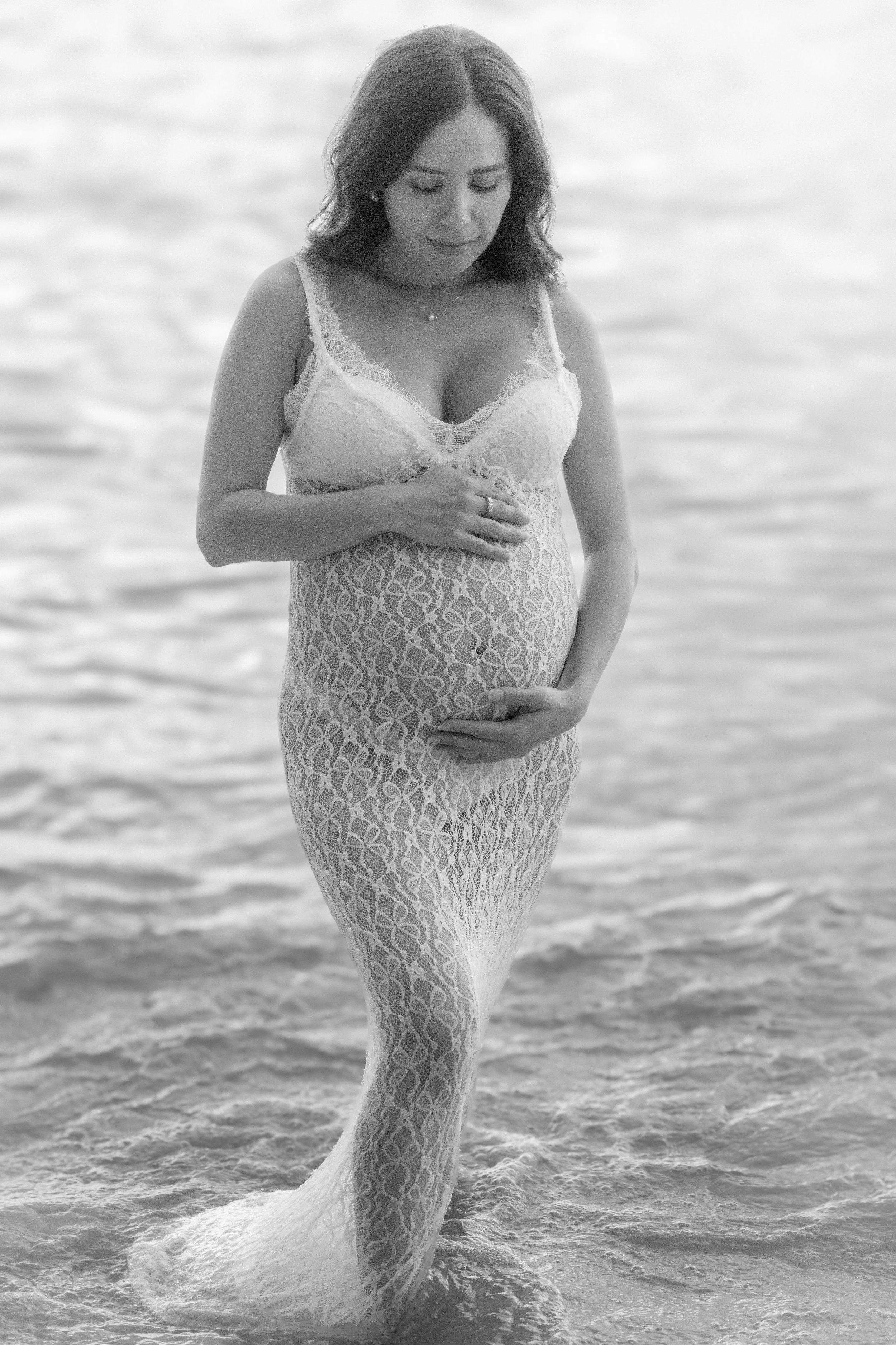 Black and white photo of a pregnant woman standing in water, wearing a lace dress, gently holding her belly, with a serene expression.