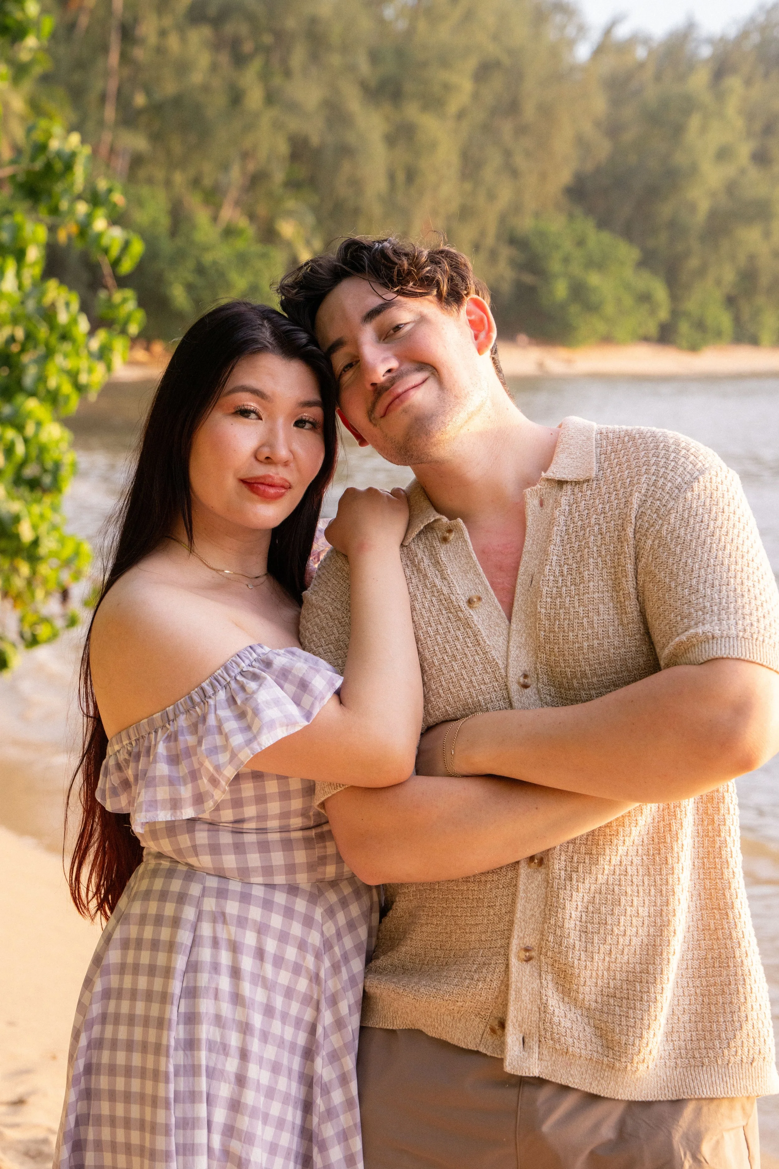 A young woman and an older man stand close together outdoors by a body of water with trees in the background. The woman has long dark hair and wears a checkered off-shoulder dress, while the man has short dark hair and wears a light-colored shirt and