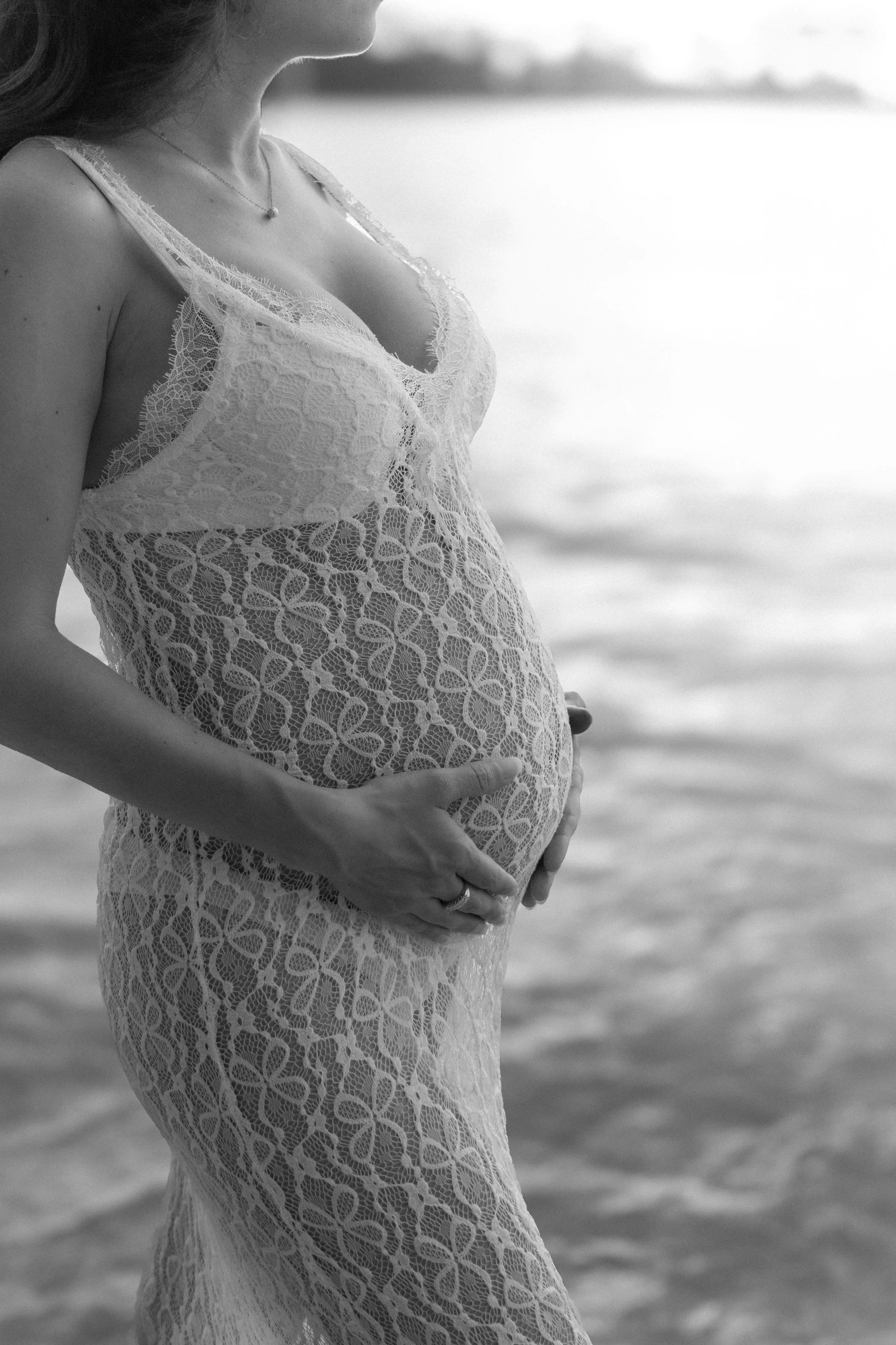 A pregnant woman in a lace dress standing on the beach during a photoshoot by isis monteux in oahu, holding her belly with both hands.