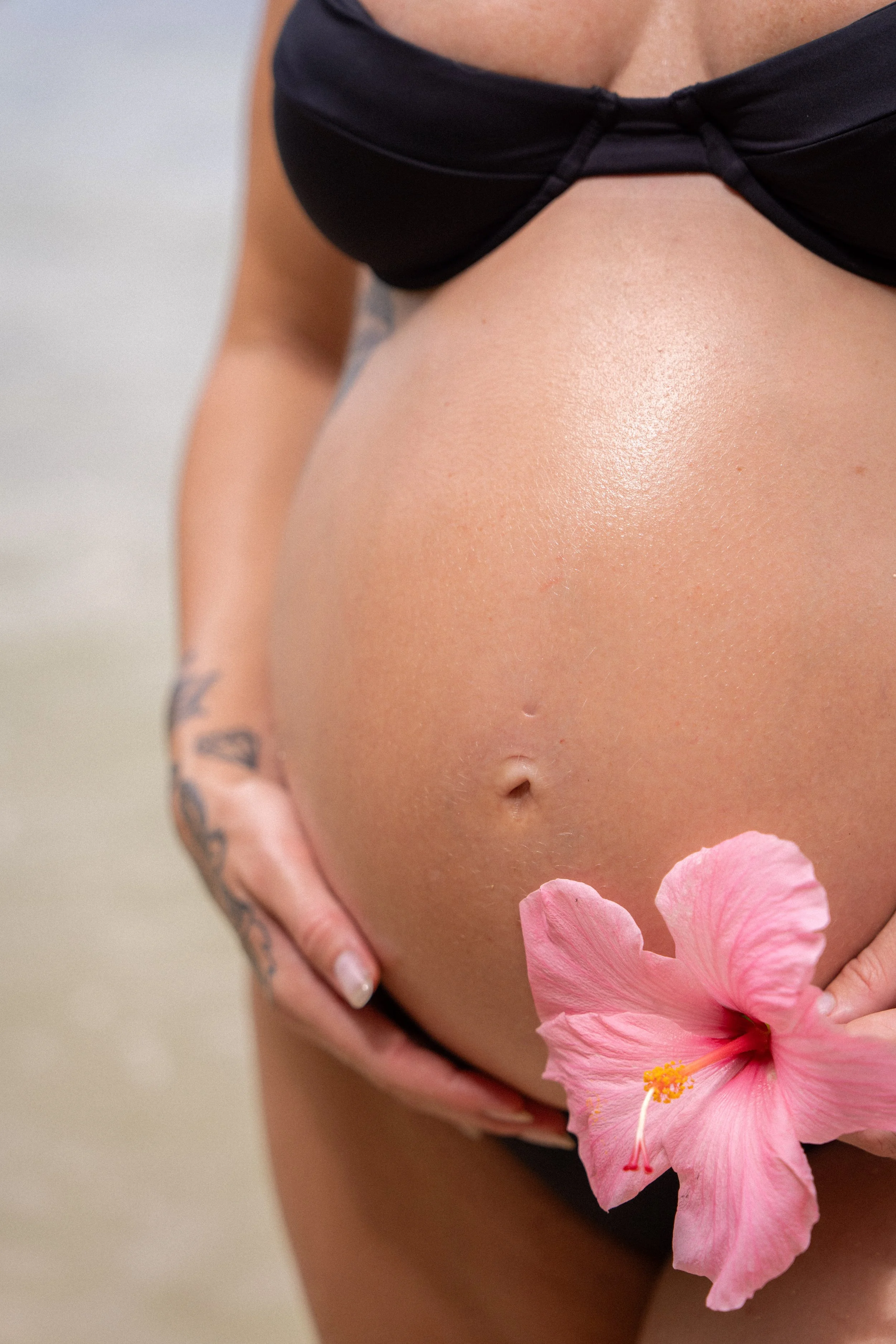 A close-up of a woman's torso, showing her toned stomach and a pink hibiscus flower held near her waist, with part of her hand visible and tattooed fingers.
