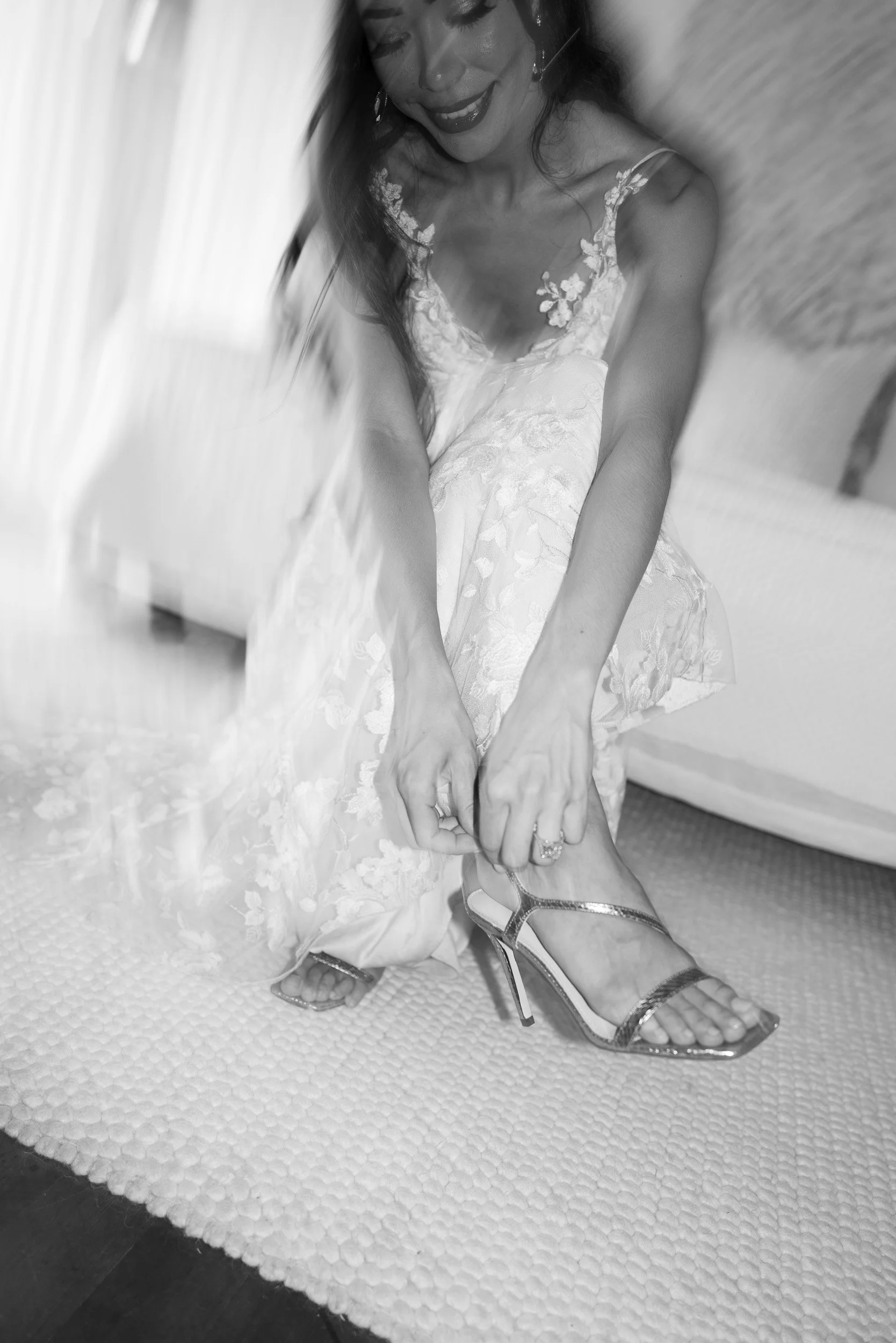 A woman in a white lace dress sitting in a cozy area, putting on high-heeled shoes on her feet, with a bright smile on her face.