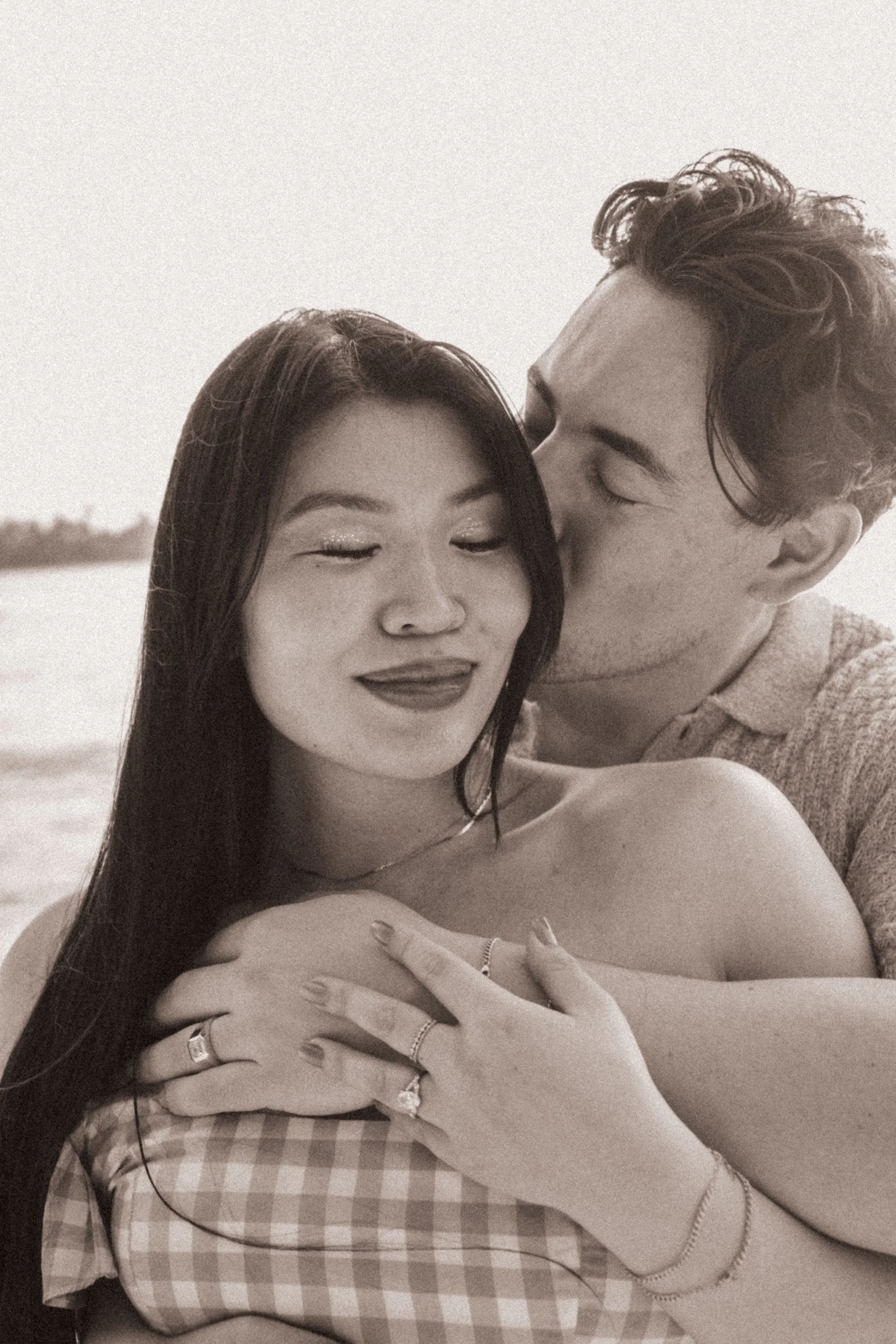 A black and white photo of a young woman and man close together outdoors. The woman has long dark hair, closed eyes, and is smiling with her lips slightly pursed. The man has wavy hair and is leaning in to kiss the woman's forehead. They are embracin