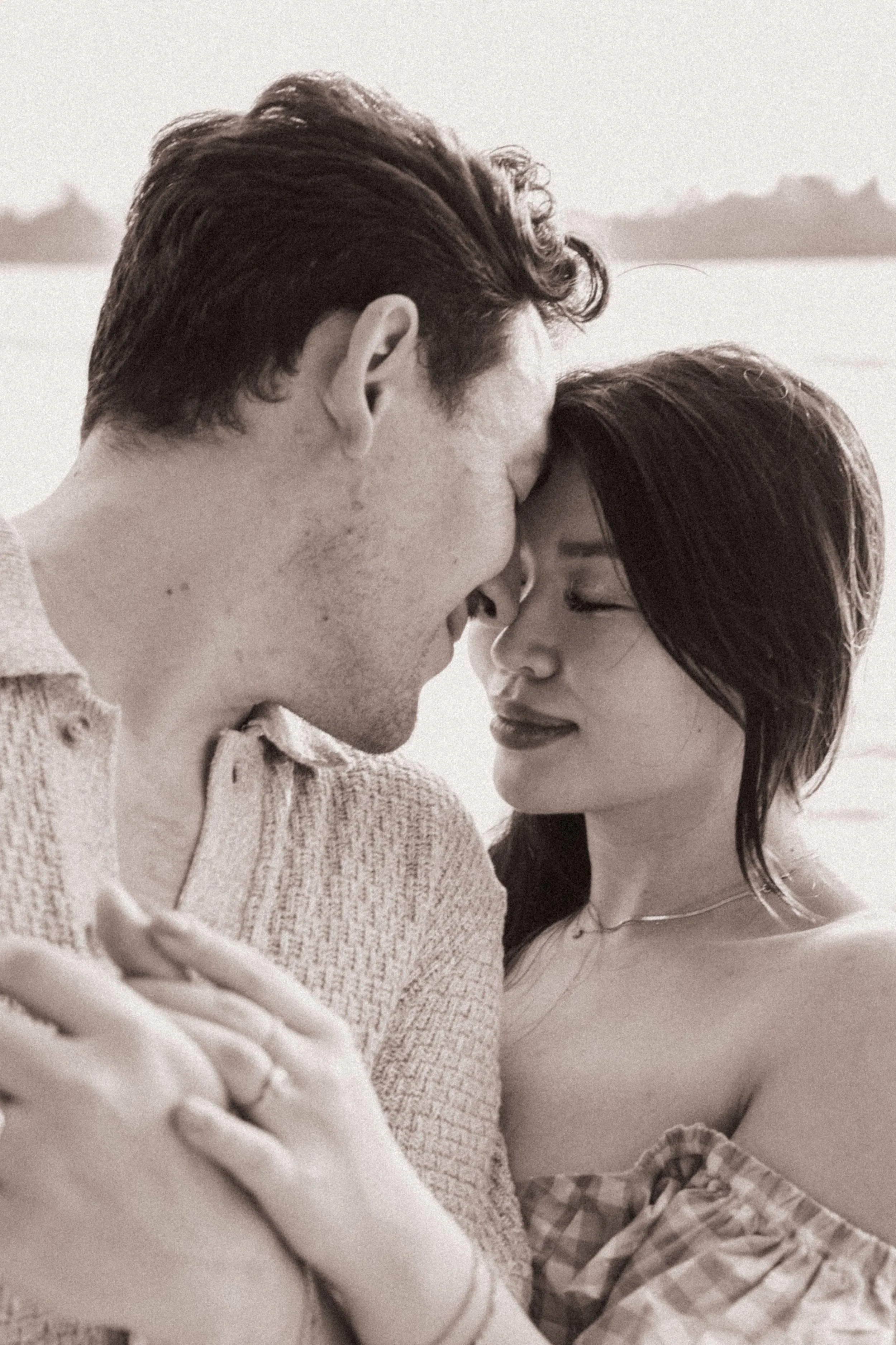 A black and white photo of a couple kissing outdoors, with the man on the left and the woman on the right. The woman's hand is resting on the man's shoulder, and she is wearing a plaid skirt and a necklace. The background appears to be a body of wate