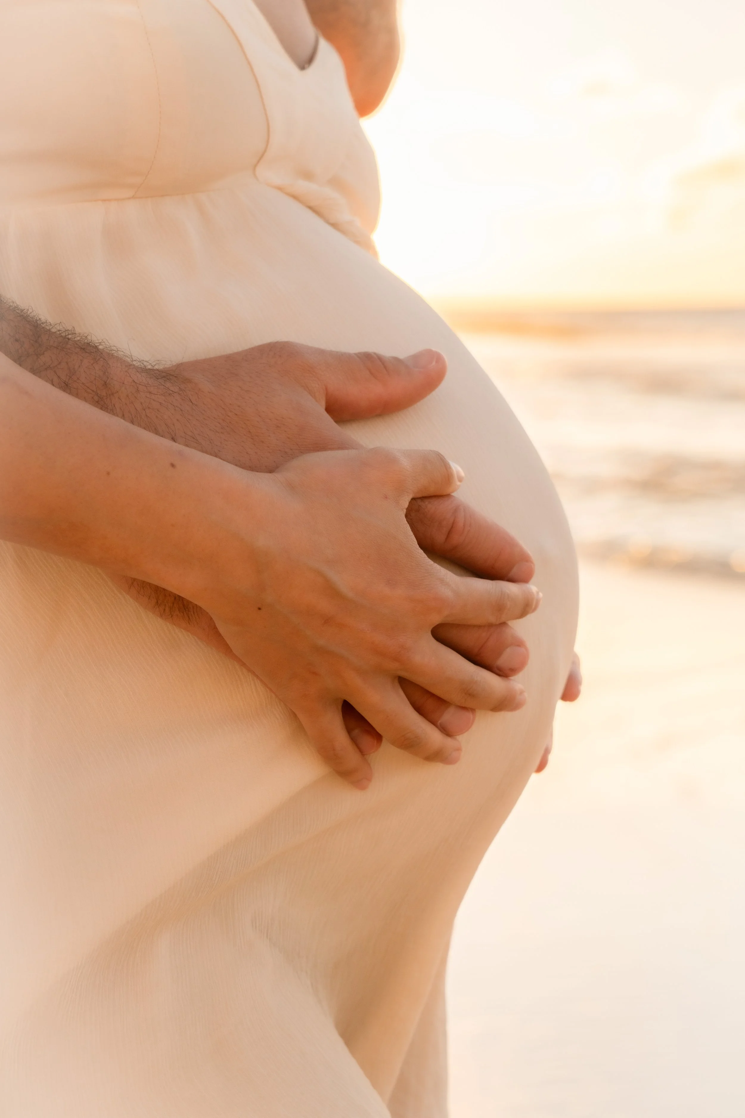 Close-up of a pregnant woman's belly with a man's hands gently holding and kissing it at a beach during sunset.