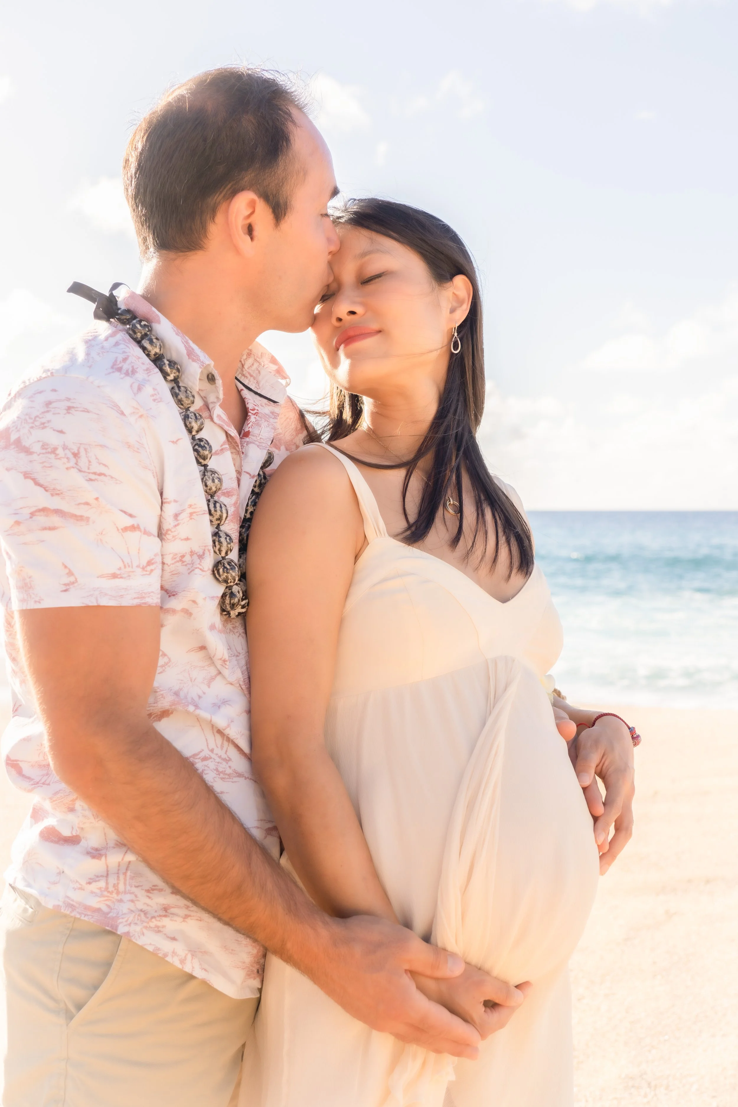 A couple on the beach on the north shore oahu, the man kissing the woman's forehead as she smiles with her eyes closed, the woman is pregnant, and they are holding her belly.