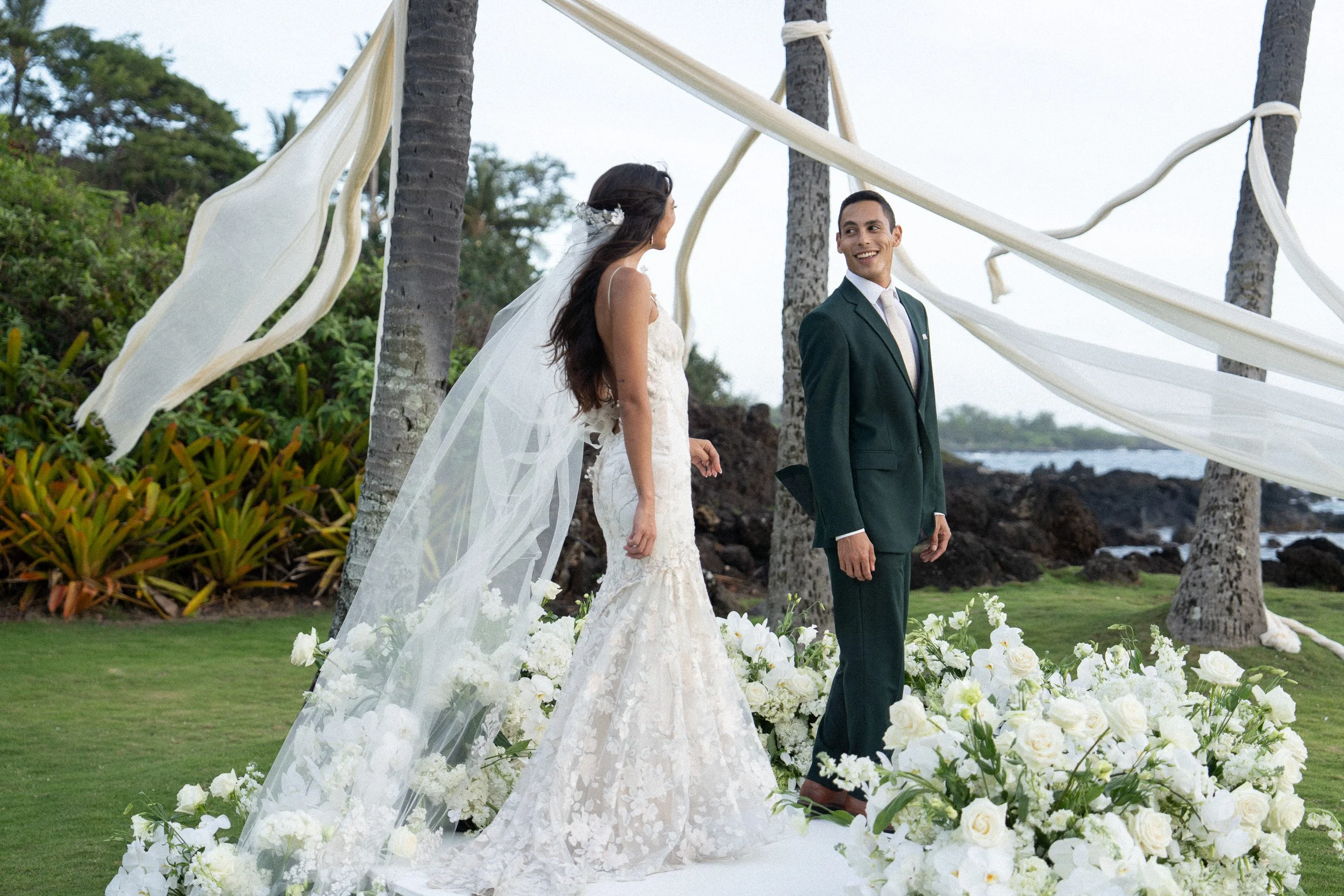 A bride and groom at an outdoor wedding ceremony on a grassy area near the beach, surrounded by trees and decorated with white flowers and flowing fabric.