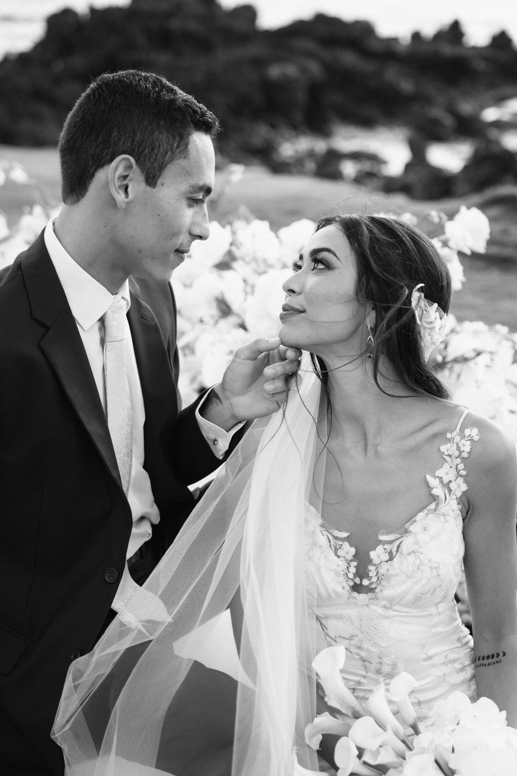 A black and white photograph of a bride and groom during their wedding ceremony. The groom is wearing a suit and tie, looking lovingly at the bride. The bride is wearing a lace wedding dress with floral embroidery, holding a bouquet of white flowers, and gazing at the groom, with a veil draped over her shoulder and hair blowing in the wind. The background features natural outdoor scenery with rocks and grass.