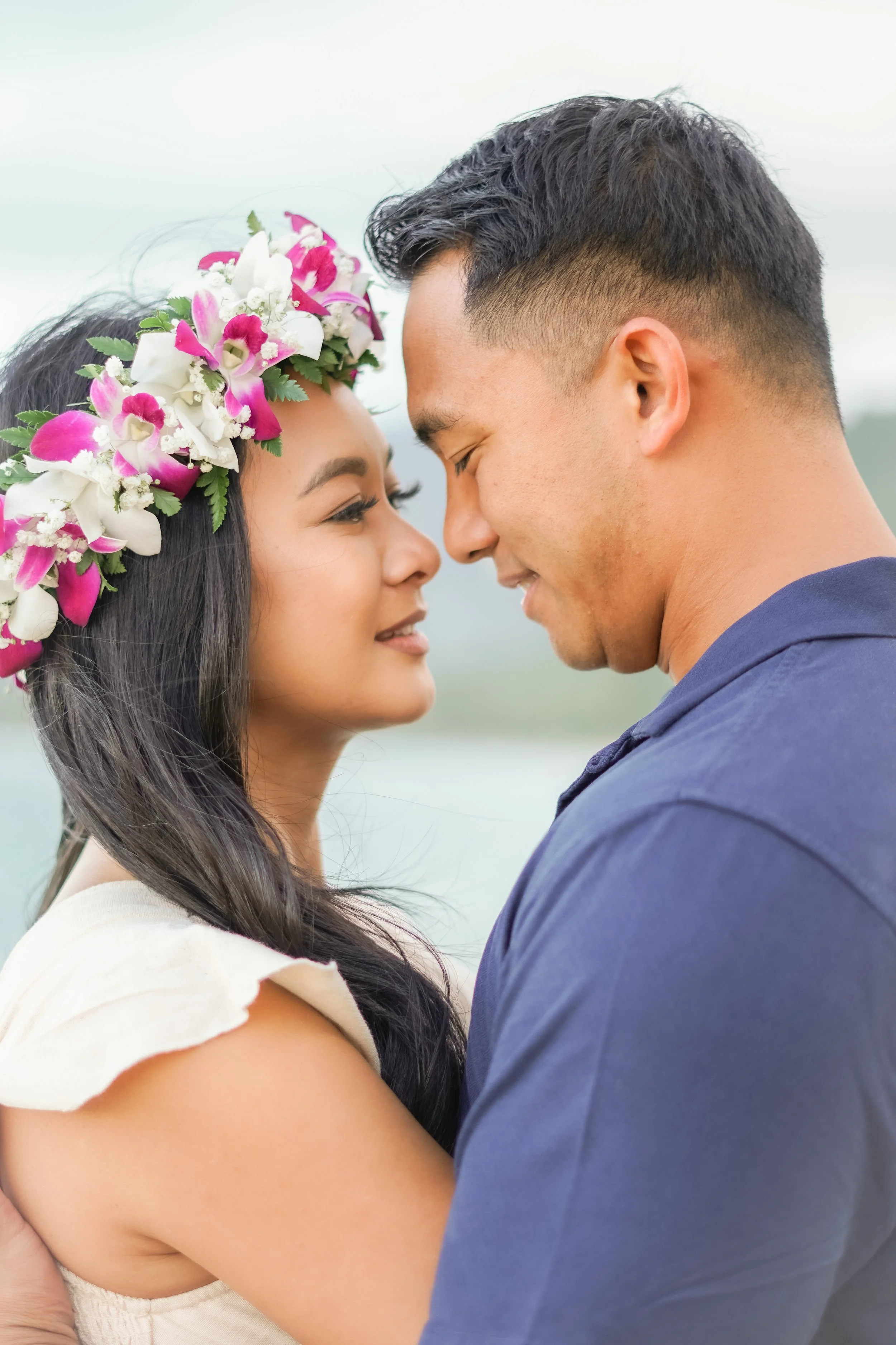 A woman wearing a flower crown with pink and white flowers and a man are standing very close with their foreheads touching and eyes closed, smiling.