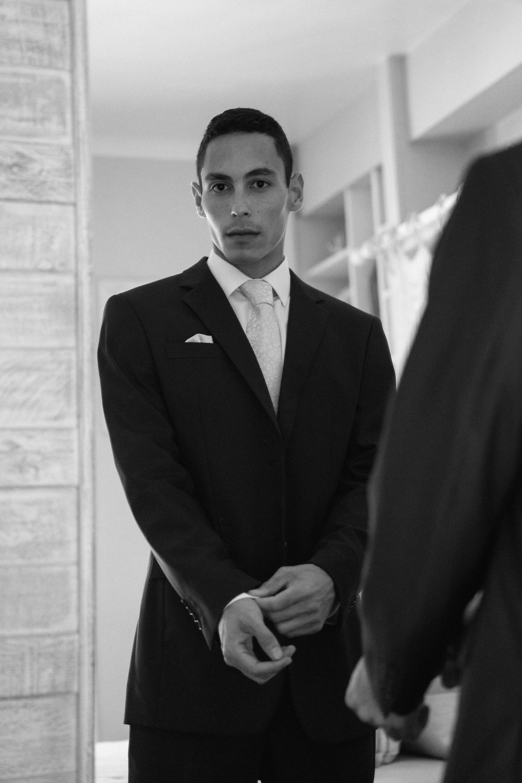 A man in a suit with a tie, standing indoors, with his hands clasped in front of him, looking serious.